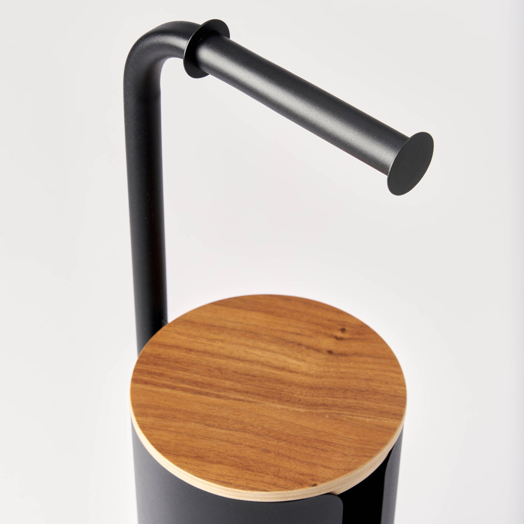 Close-up of the curved black steel arm and round wood lid on the Apto Aloft Toilet Paper Stand with Storage, shown on a white background