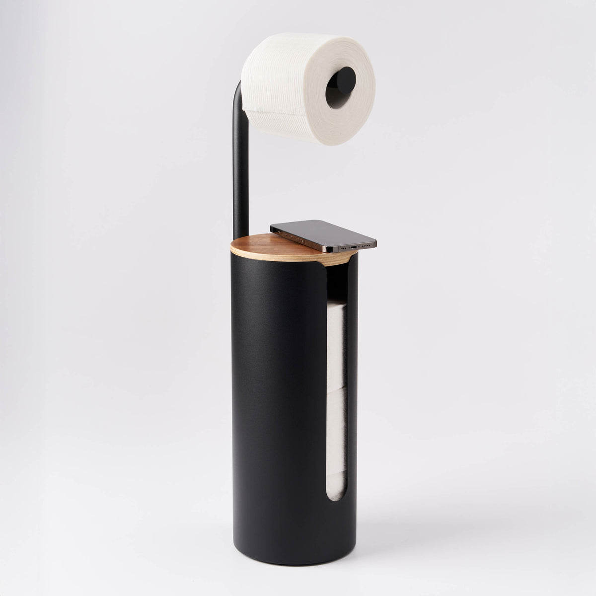 Apto Aloft Toilet Paper Stand with Storage in black with a phone resting on the wood lid and a roll on the arm, shown on a white background