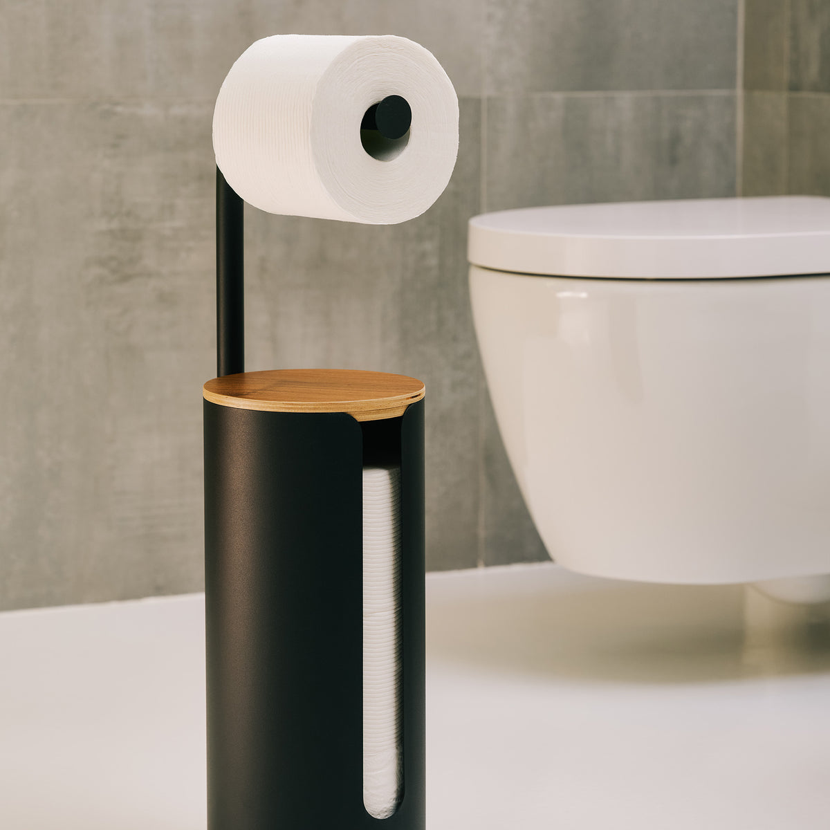 Apto Aloft Toilet Paper Stand with Storage in black steel and wood, holding a roll beside a wall-mounted toilet in a gray tiled bathroom