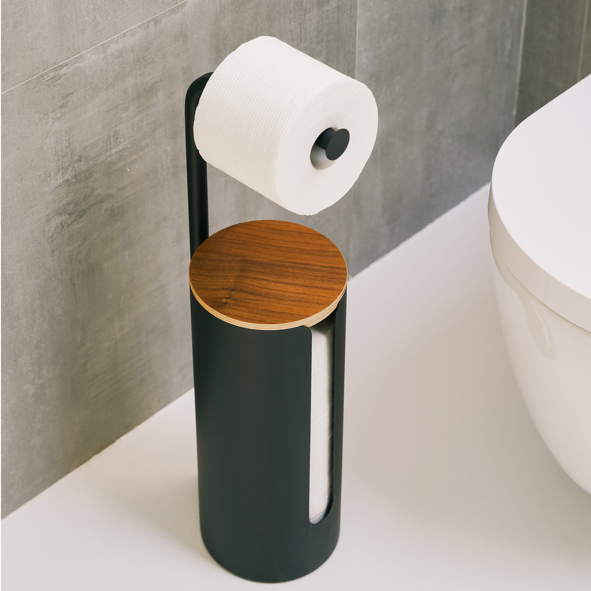 Angled view of the Apto Aloft Toilet Paper Stand with Storage in black, showing the round wood lid and a roll on the arm next to a toilet