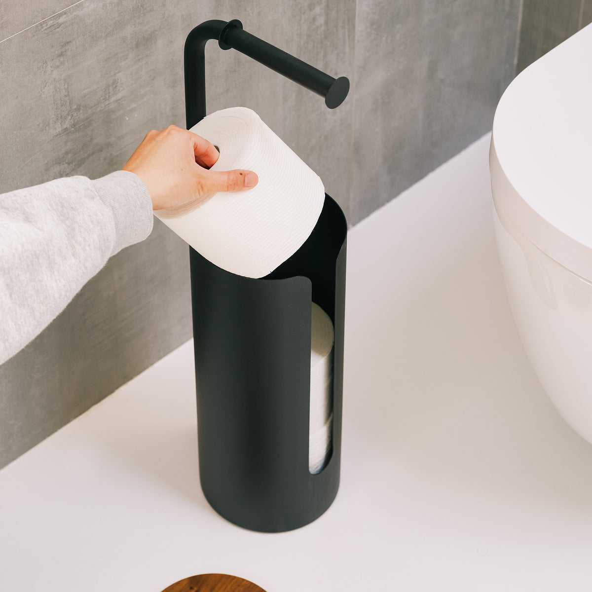 Hand dropping a spare roll into the open canister of the Apto Aloft Toilet Paper Stand with Storage in black steel, wood lid set aside