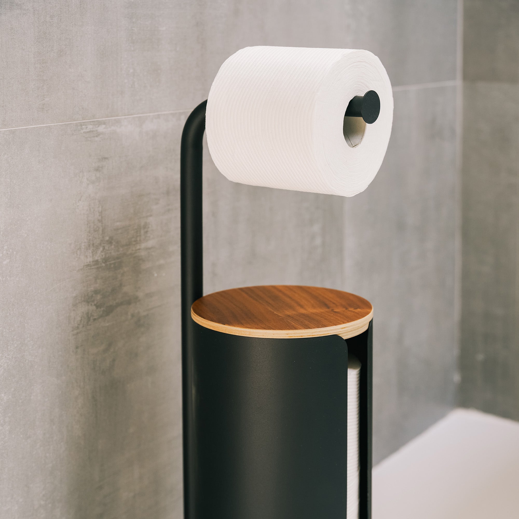 Close-up of the Apto Aloft Toilet Paper Stand with Storage in black steel, with its wood lid and a roll on the arm against a gray tile wall