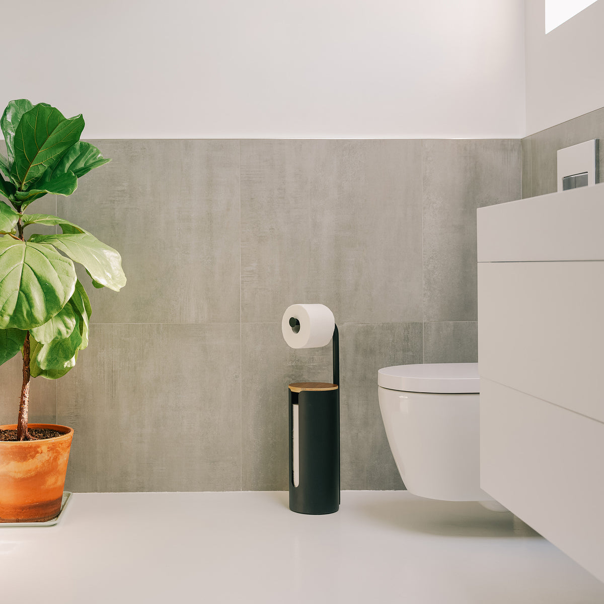Apto Aloft Toilet Paper Stand with Storage in black standing next to the toilet in a bright bathroom with a white vanity and potted plant