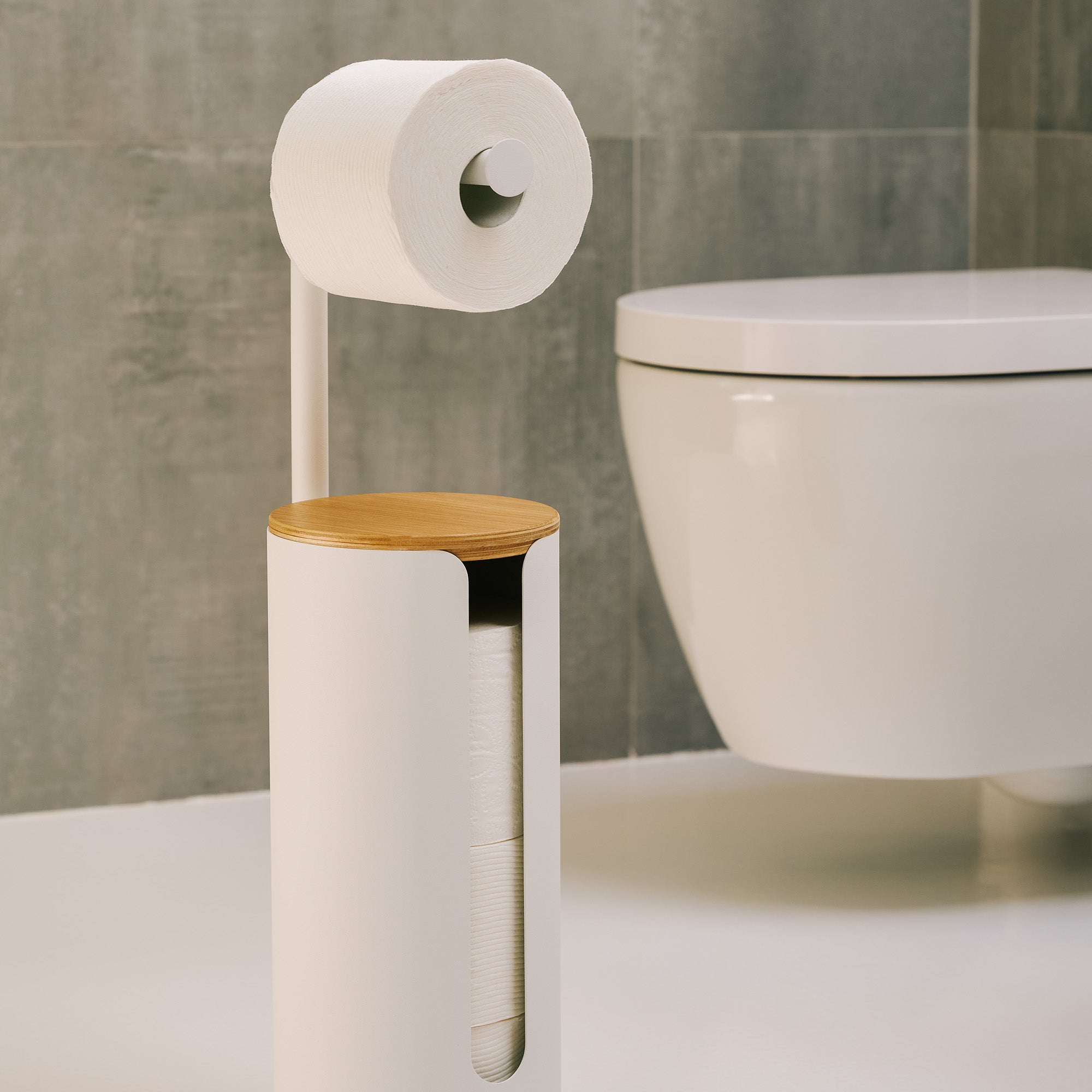 Apto Aloft Toilet Paper Stand with Storage in white steel and wood, holding a roll beside a wall-mounted toilet in a gray tiled bathroom