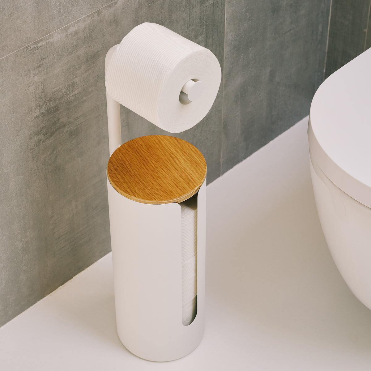 Angled view of the Apto Aloft Toilet Paper Stand with Storage in white, showing the round wood lid and a roll on the arm next to a toilet