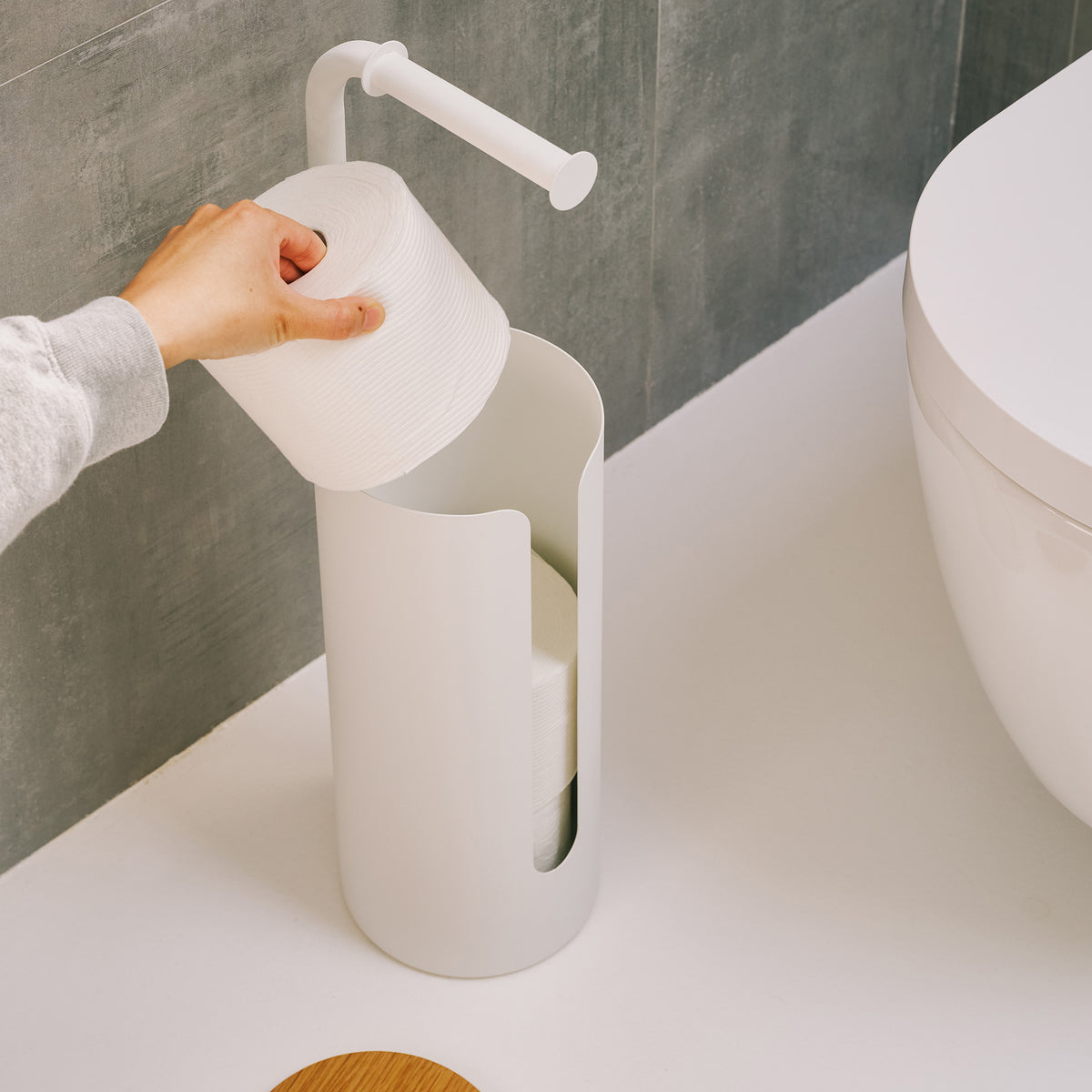 Hand dropping a spare roll into the open canister of the Apto Aloft Toilet Paper Stand with Storage in white steel, wood lid set aside