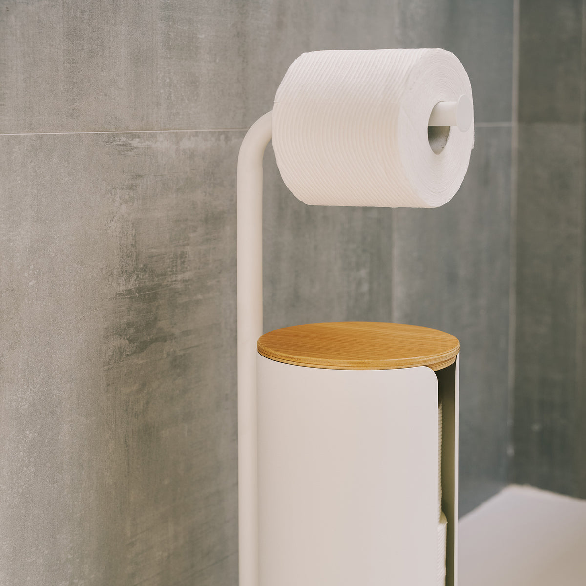 Close-up of the Apto Aloft Toilet Paper Stand with Storage in white steel, with its wood lid and a roll on the arm against a gray tile wall