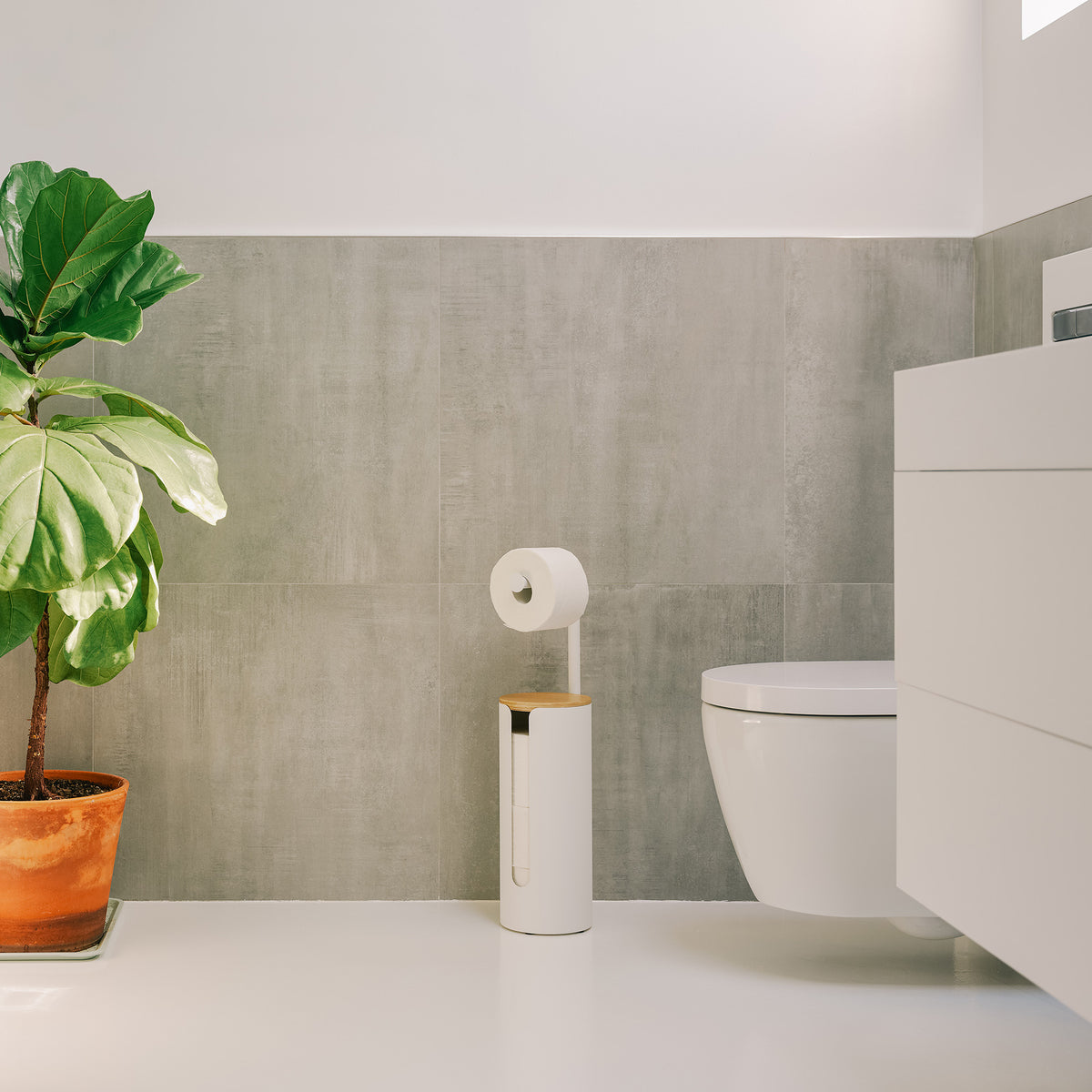 Apto Aloft Toilet Paper Stand with Storage in white standing next to the toilet in a bright bathroom with a white vanity and potted plant