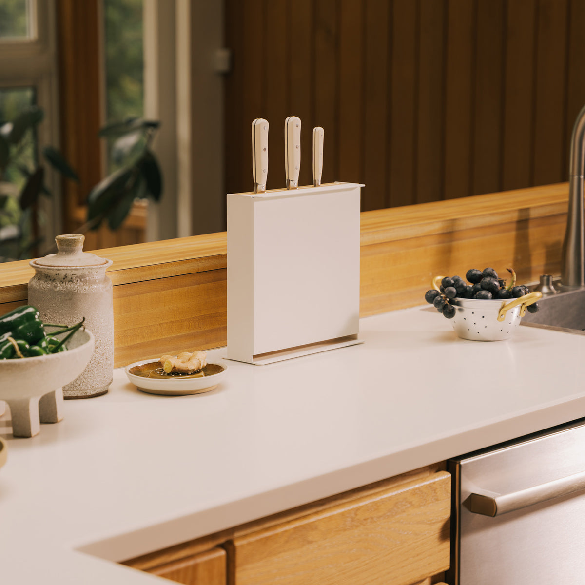 Apto Slot Knife Stand and Cutting Board in white steel holding three knives on a kitchen counter with warm wood cabinets behind it