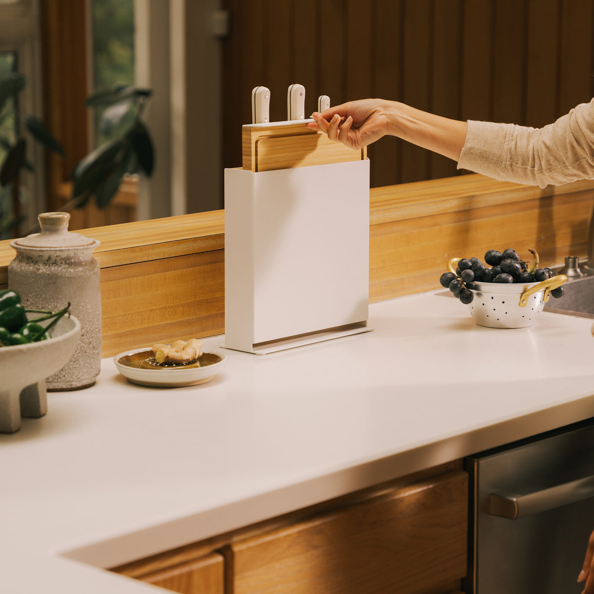 Hand sliding the bamboo board out of the Apto Slot Knife Stand and Cutting Board, which holds three knives on a white kitchen counter