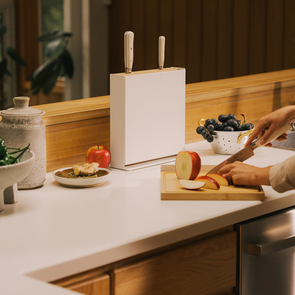 Person slicing an apple on the bamboo board from the Apto Slot Knife Stand and Cutting Board, with the stand and knives on the counter