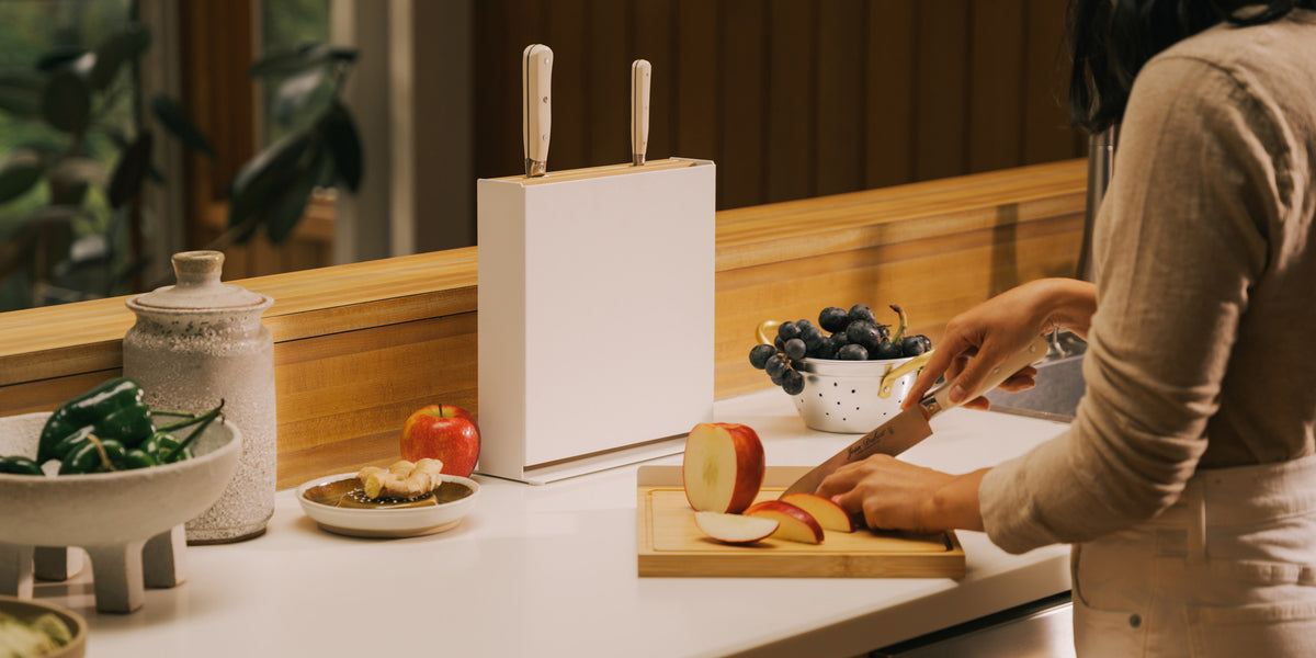 Person slicing an apple on the bamboo board from the Apto Slot Knife Stand and Cutting Board, with the stand and knives on the counter