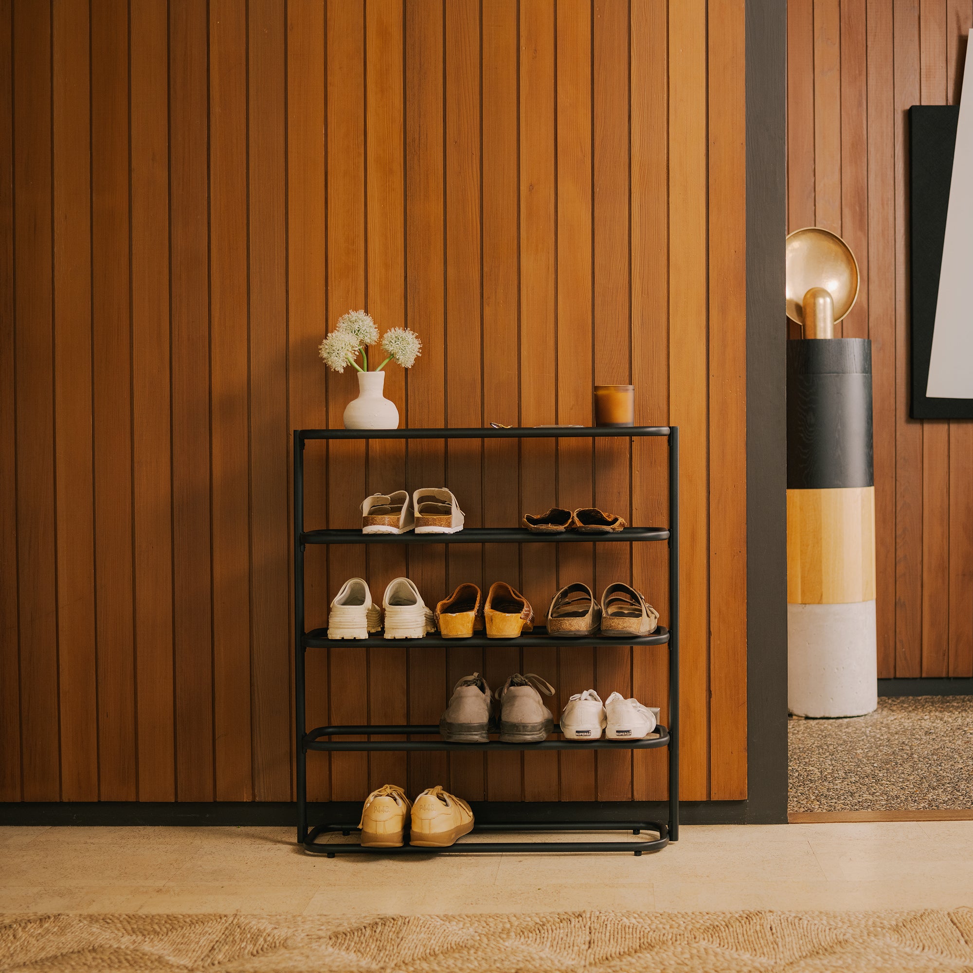 Black 4-tier metal shoe rack with tabletop against a wood-paneled wall, holding 8 pairs of shoes, topped with a vase and candle.