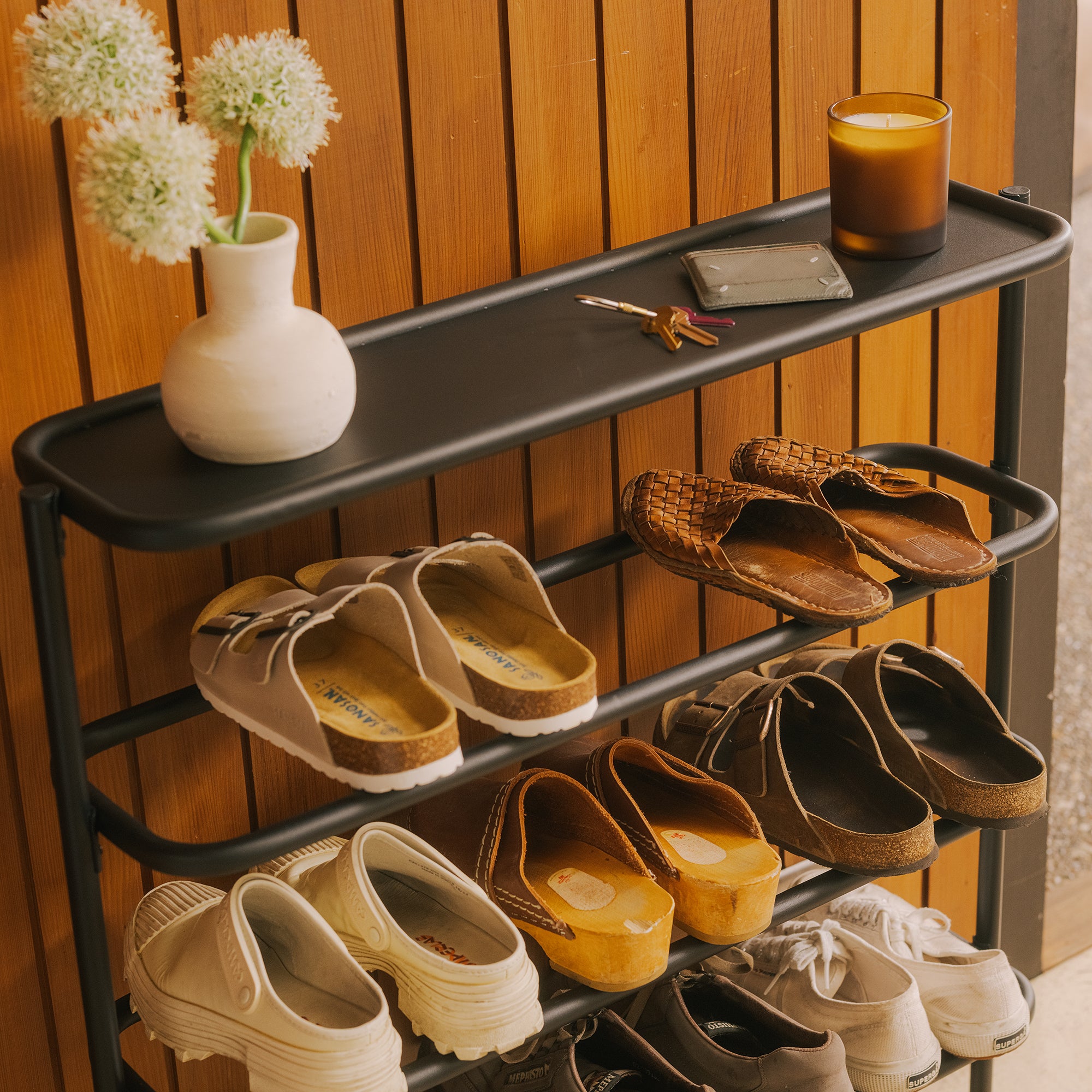 Black metal entryway shoe rack with flat top shelf holding flowers, candle, and keys, filled with sandals and clogs