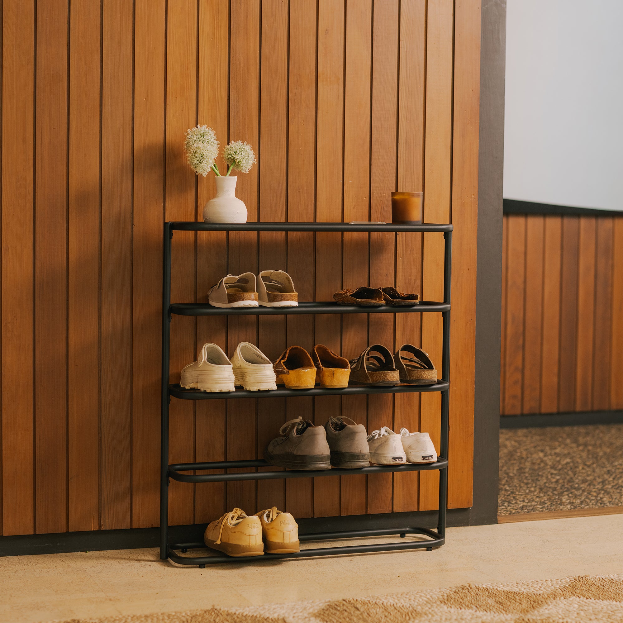 Black 4-tier metal shoe rack with tabletop against a wood-paneled wall, holding 8 pairs of shoes, topped with a vase and candle.