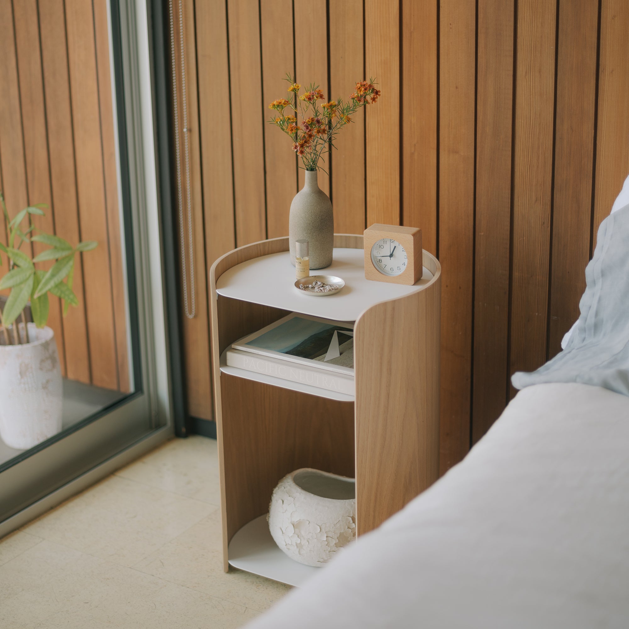 Modern ash wood and white bedside table with open shelves, styled with ceramic vase, clock, and books in minimalist bedroom