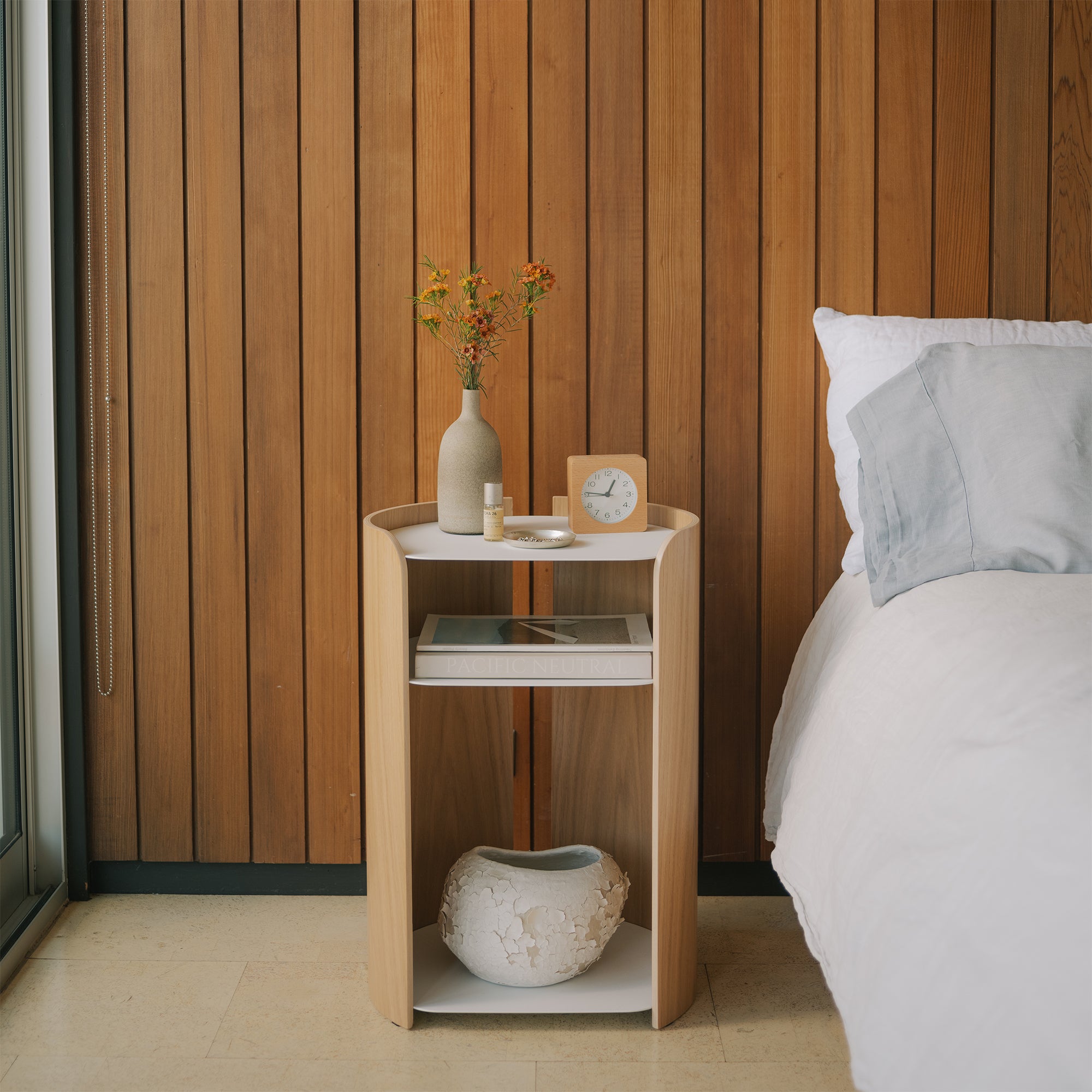 Scandinavian oak and white nightstand with curved edges and open shelves next to bed, styled with vase and clock