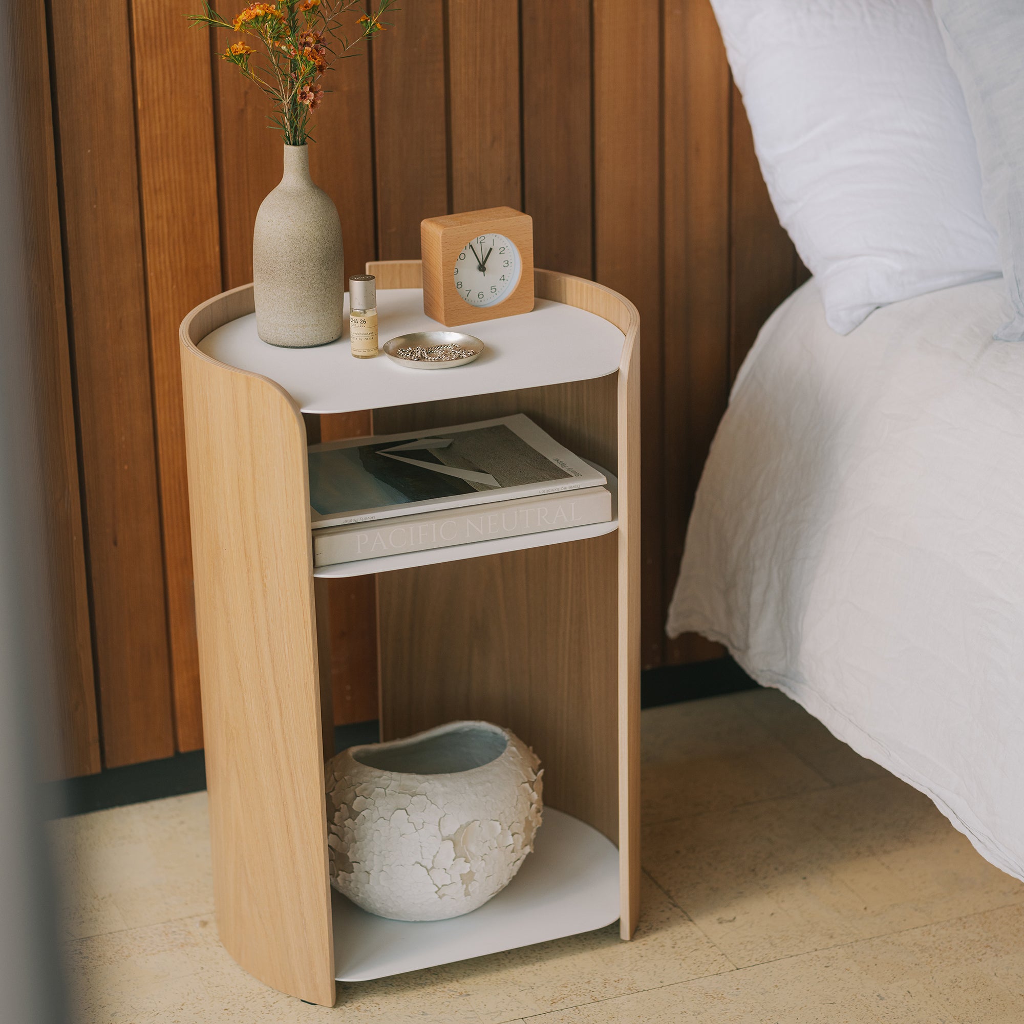 Ash and white nightstand with three open shelves styled with vase, Le Labo Matcha 26 perfume, books, and ceramic sculpture beside white bed