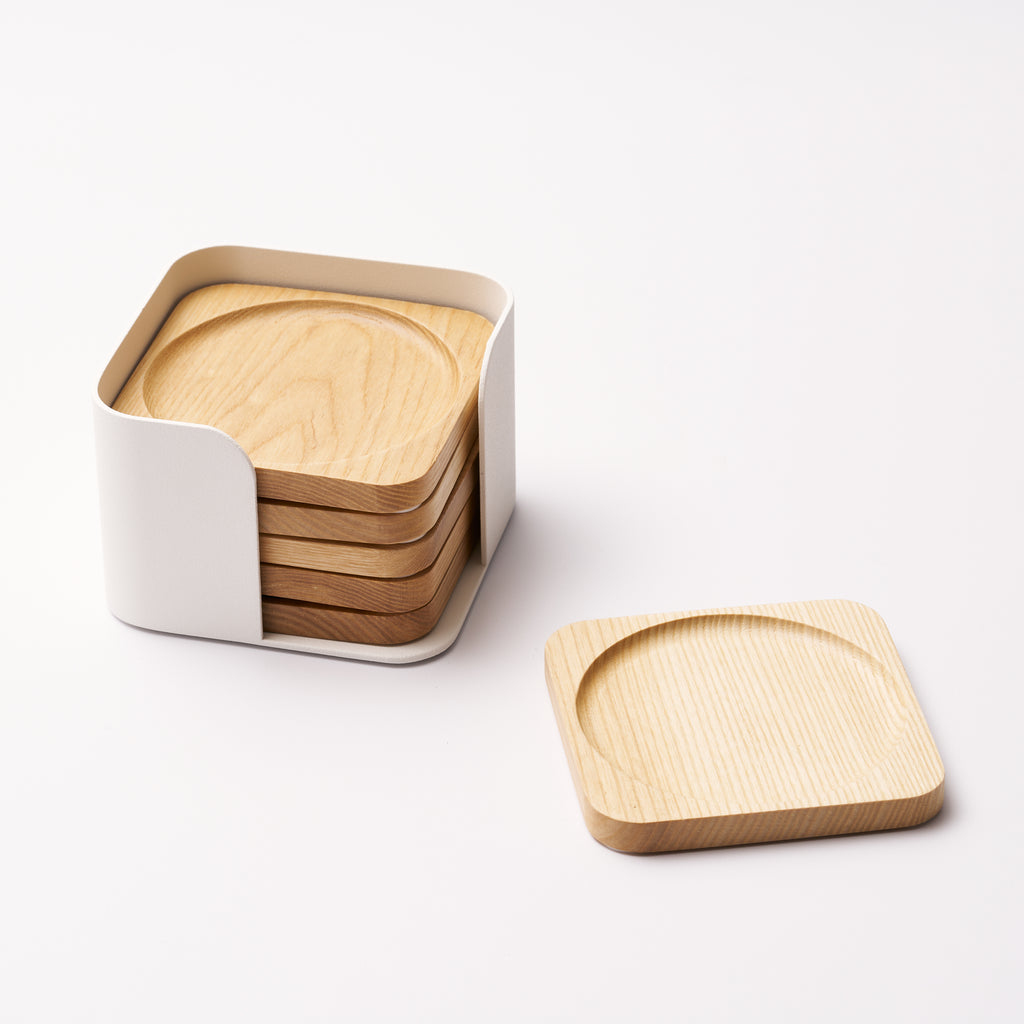 Five natural wood coasters held by a white steel organizer and one coaster displayed on its own on a white background. Designed by Apto. 