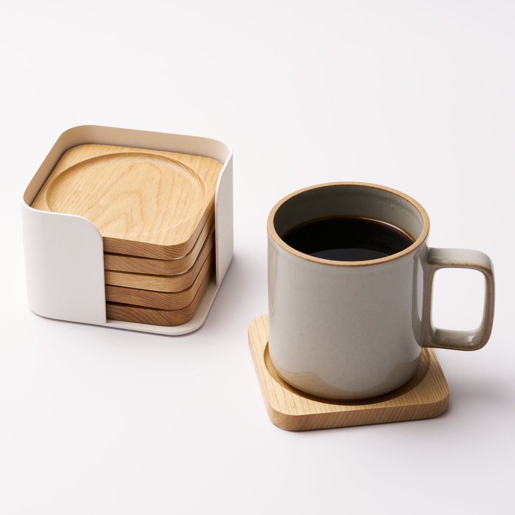 Five natural wood coasters held by a white steel organizer and one coaster displayed holding a coffee mug on a white background. Designed by Apto. 