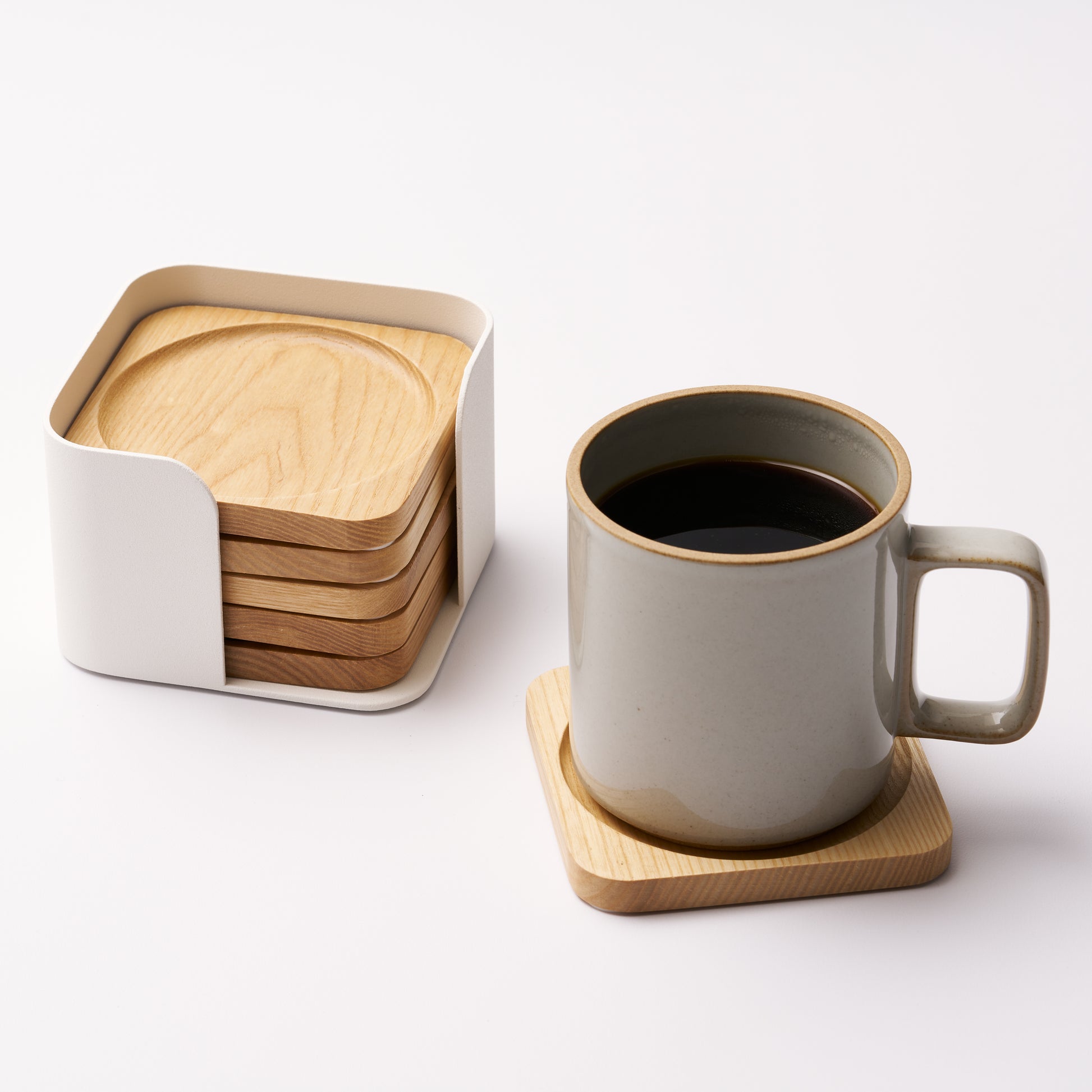 Five natural wood coasters held by a white steel organizer and one coaster displayed holding a coffee mug on a white background. Designed by Apto. 