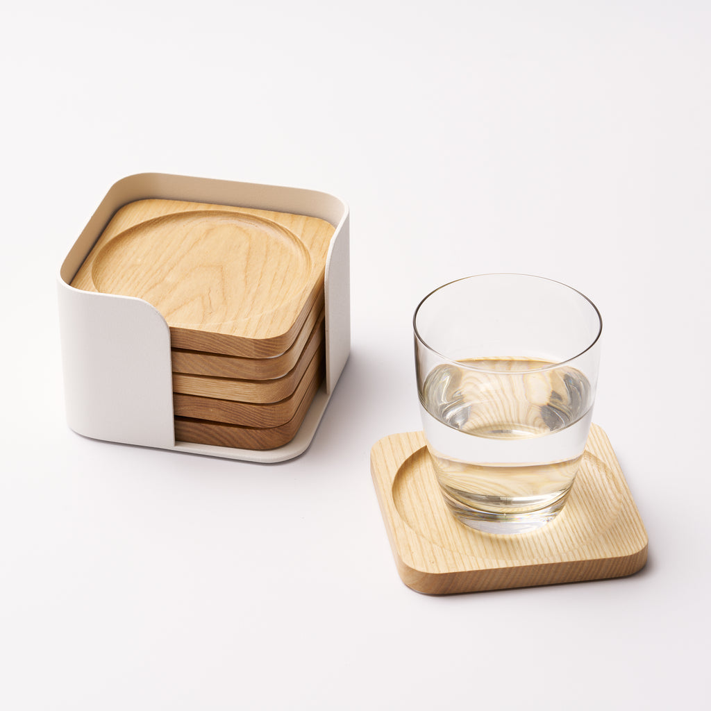 Five natural wood coasters held by a white steel organizer and one coaster displayed holding a glass of water in a clear glass on a white background. Designed by Apto. 