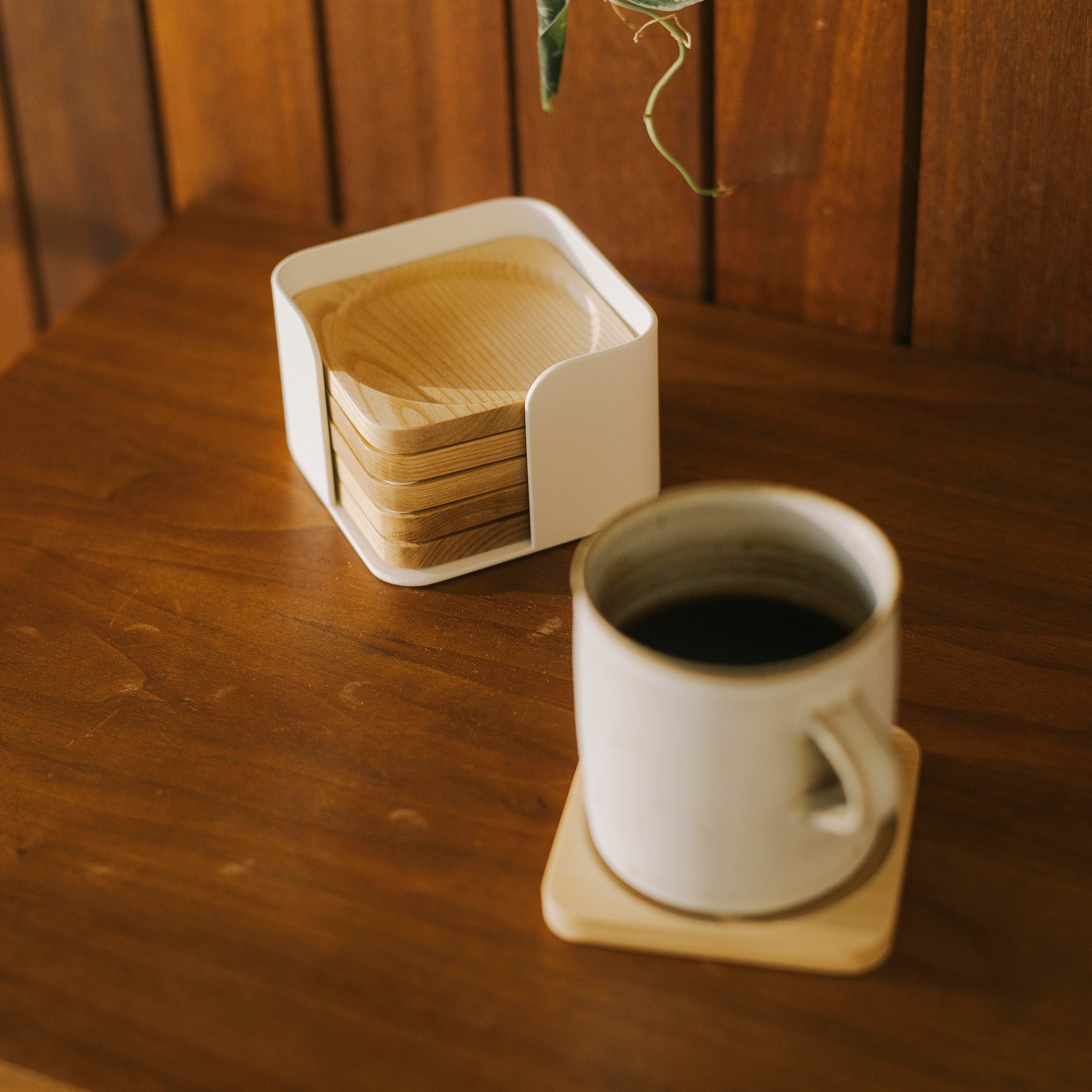 Contour Coaster Set shown with five out of six natural wood coasters in a white steel container and one coaster placed under a ceramic coffee mug in a dark wood living room.