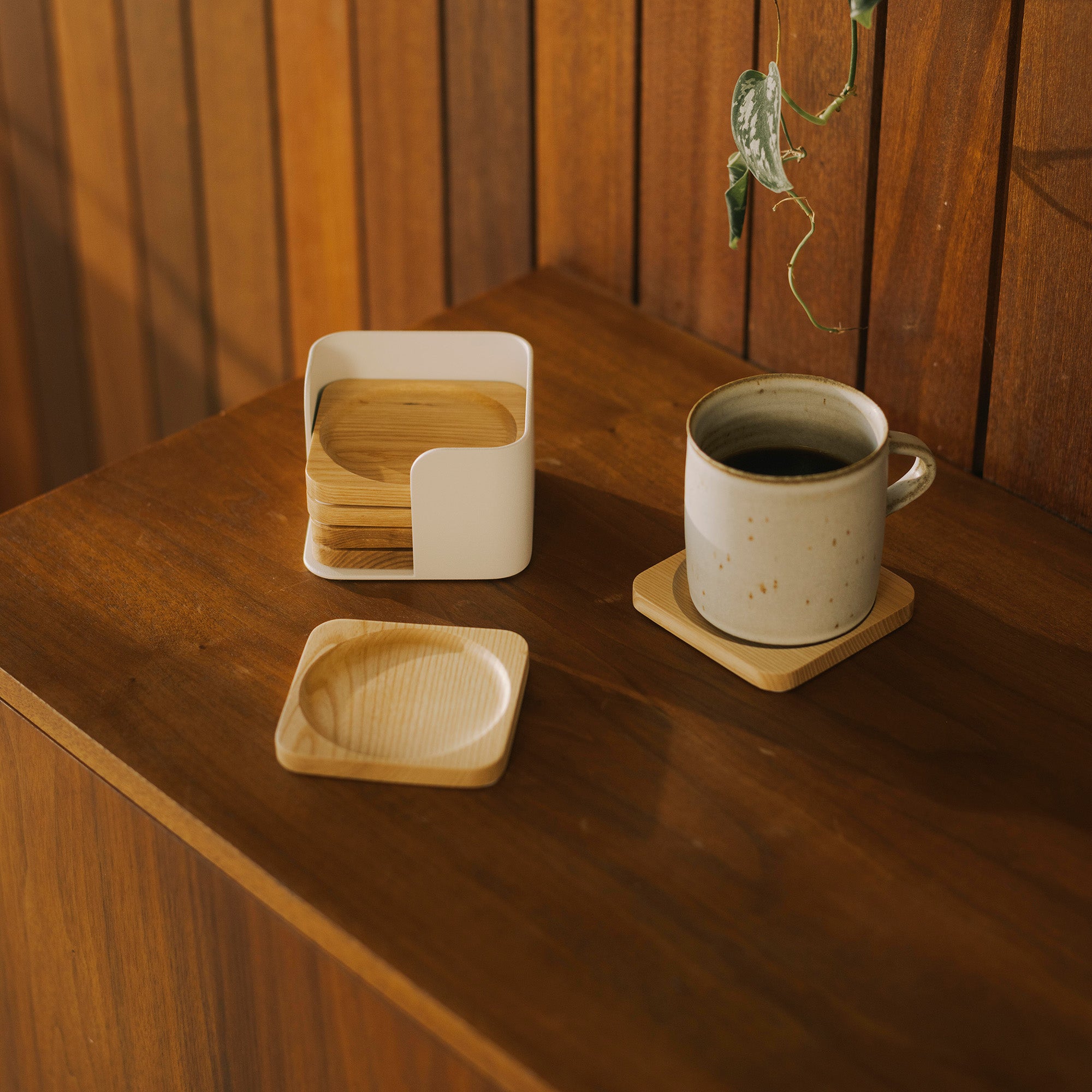 Contour Coaster Set shown holding four out of six natural wood coasters in the white steel container and one coaster placed under a ceramic coffee mug and one on the coffee table to show a center divet that holds drinks in place. 