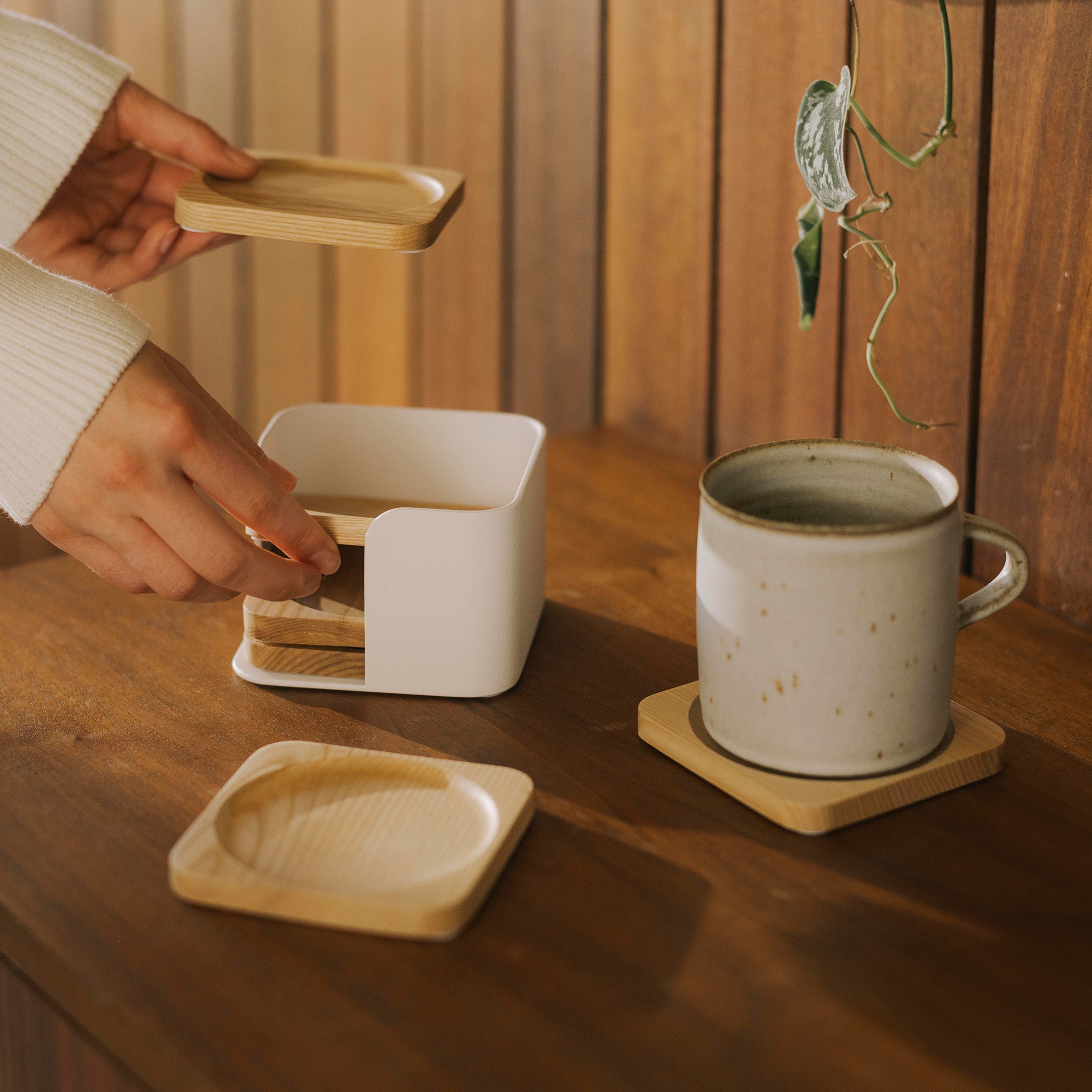 Woman organizes wood coasters inside the white steel organizing tray. One coaster is being used for a coffee mug and one is displayed, showing the center divet for drinks. 
