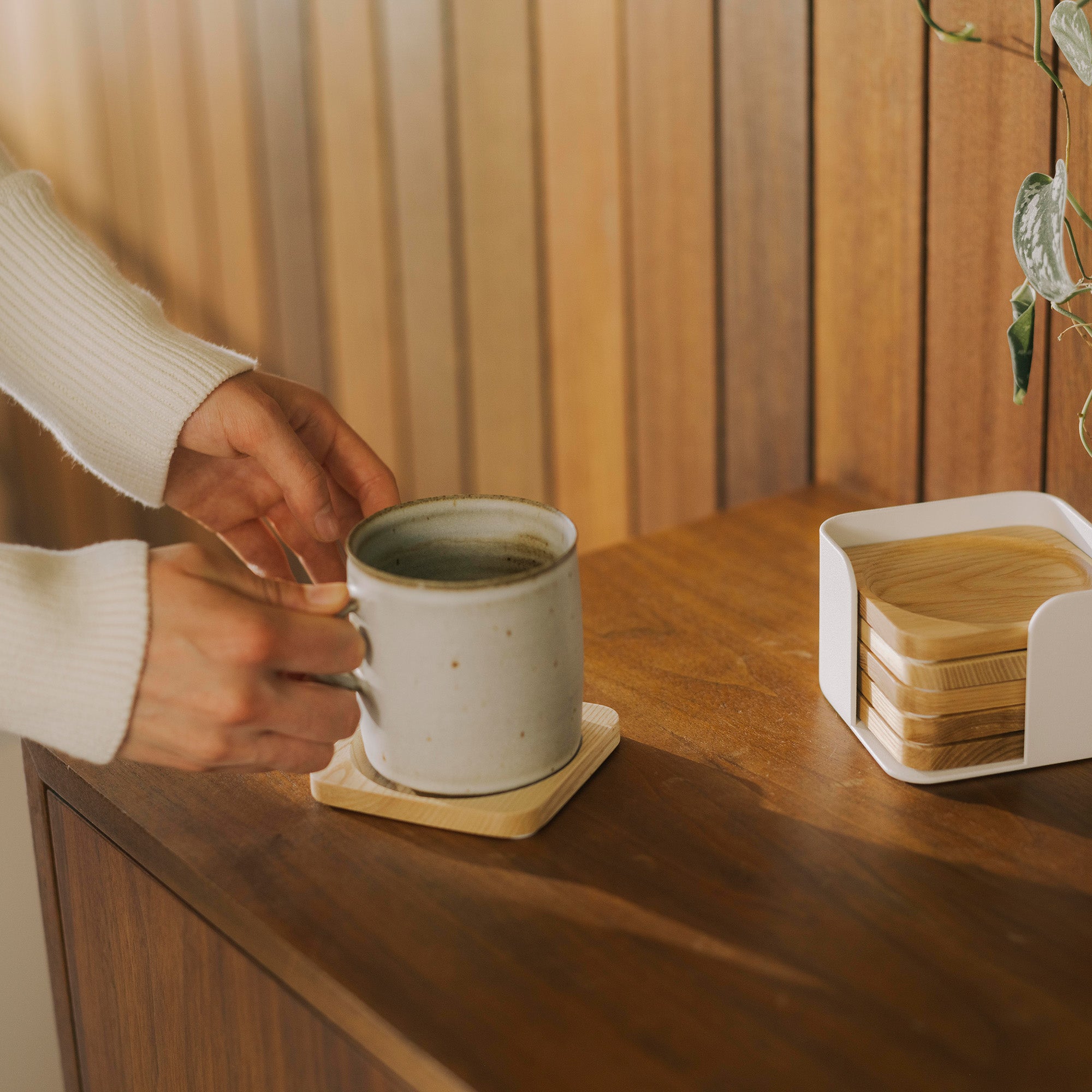 Hand picks up coffee mug from natural wood coaster in a living room furnished with dark wood. Coaster set designed by Apto. 