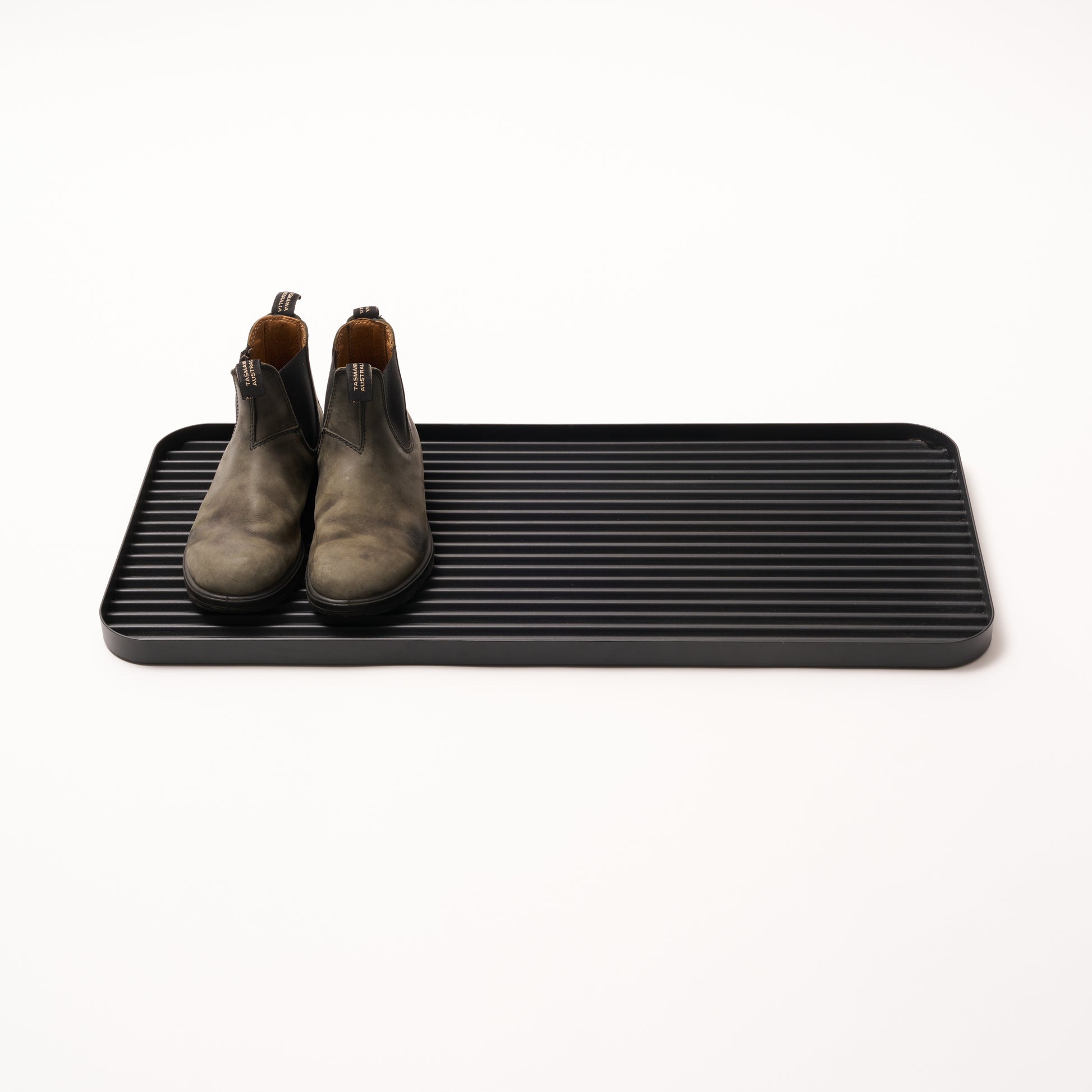 Black steel boot tray with ridged bottom holding a pair of boots with a white backdrop. Designed by Apto. 
