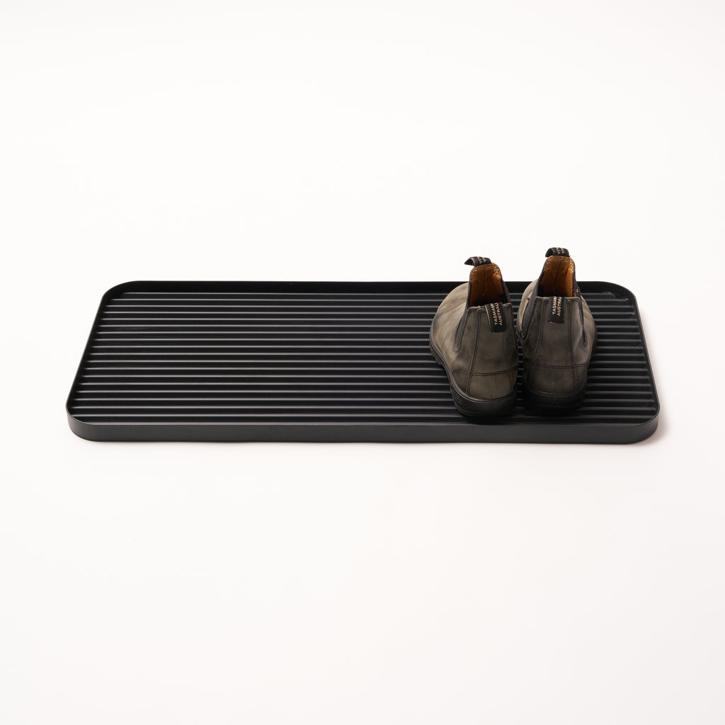 Black steel boot tray with ridged bottom holding a pair of boots with a white backdrop. Designed by Apto. 