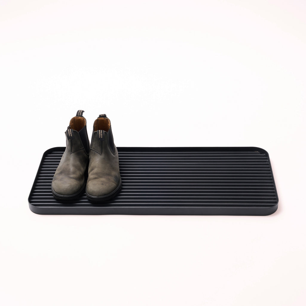 Black steel boot tray with ridged bottom holding a pair of boots with a white backdrop. Designed by Apto. 