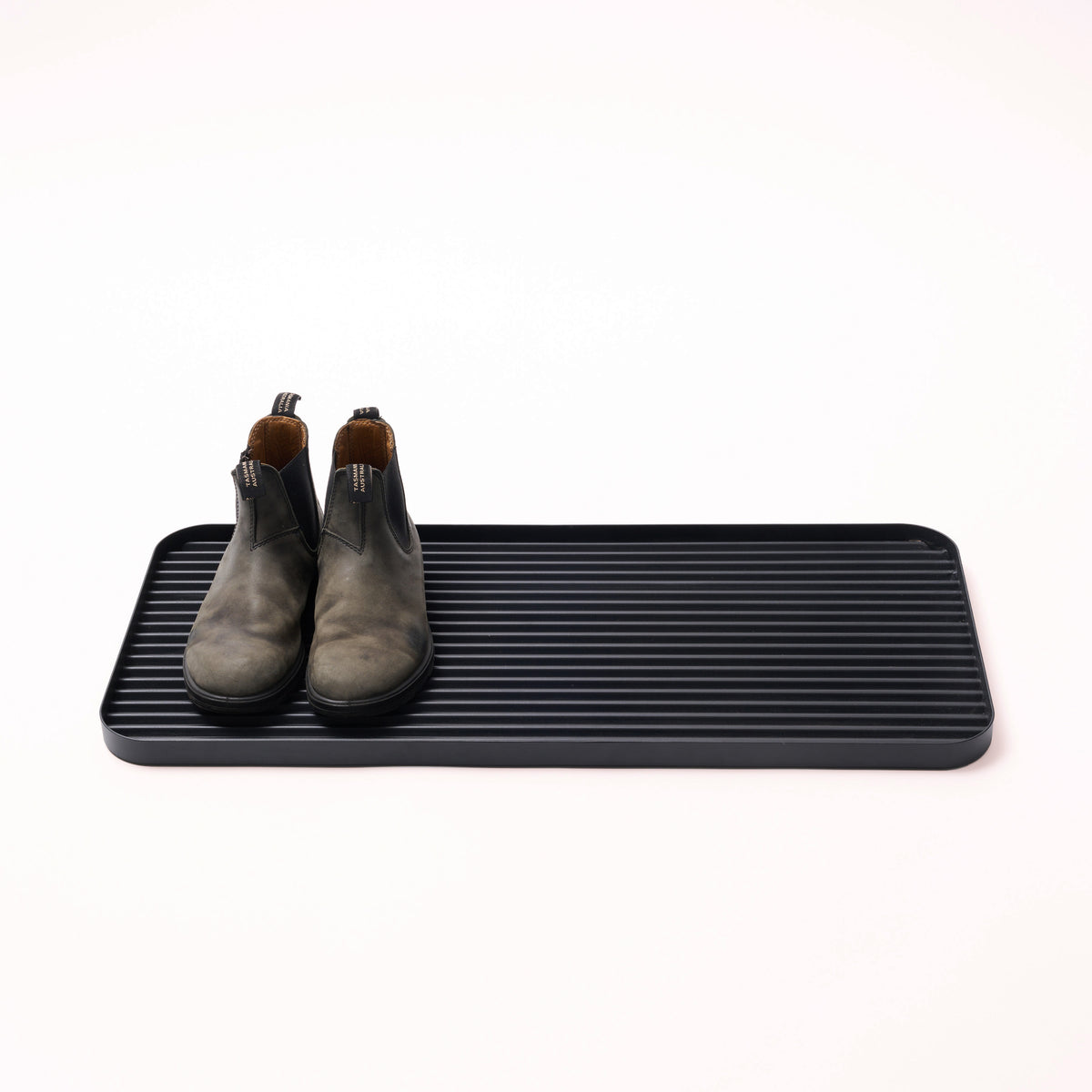 Black steel boot tray with ridged bottom holding a pair of boots with a white backdrop. Designed by Apto. 