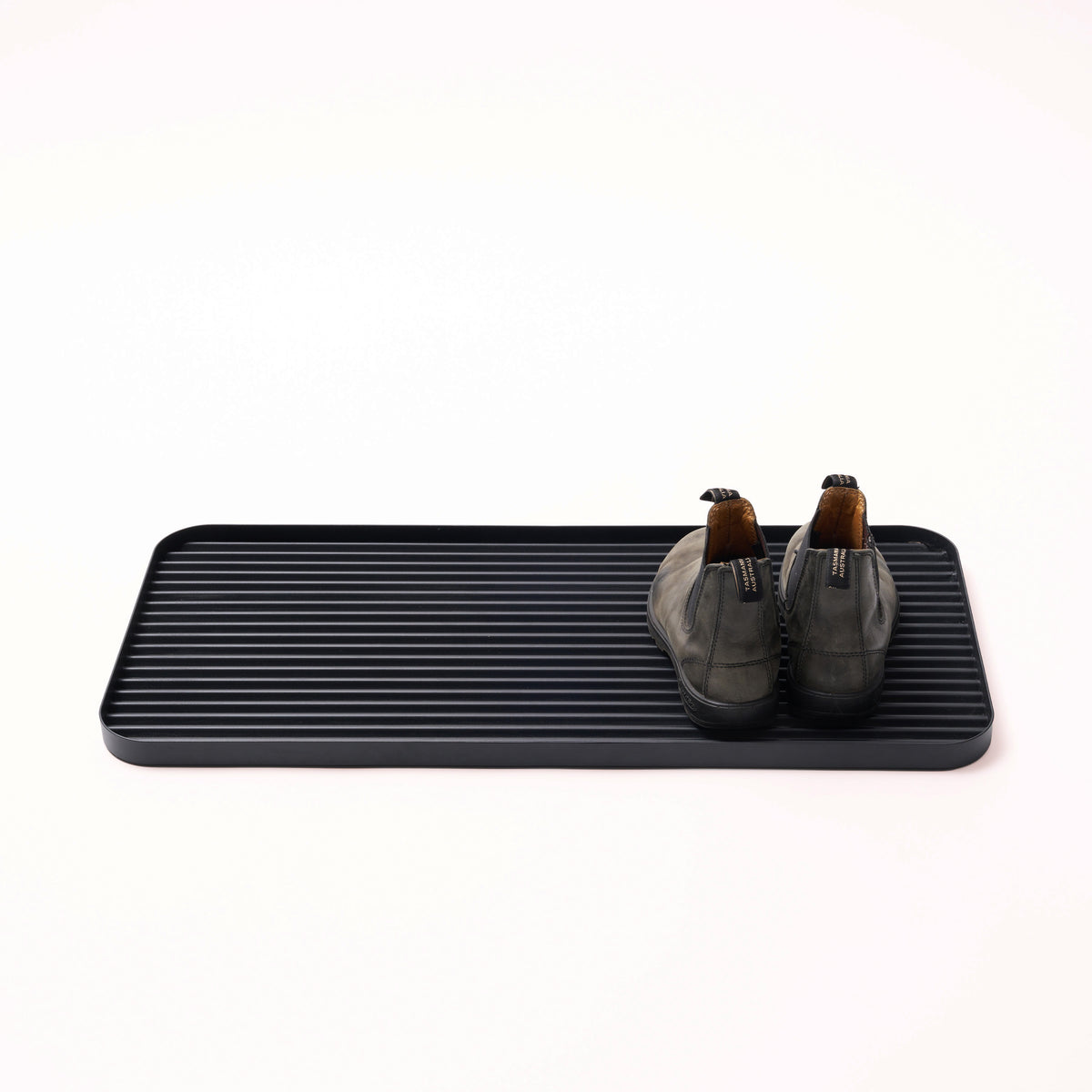 Black steel boot tray with ridged bottom holding a pair of boots with a white backdrop. Designed by Apto. 