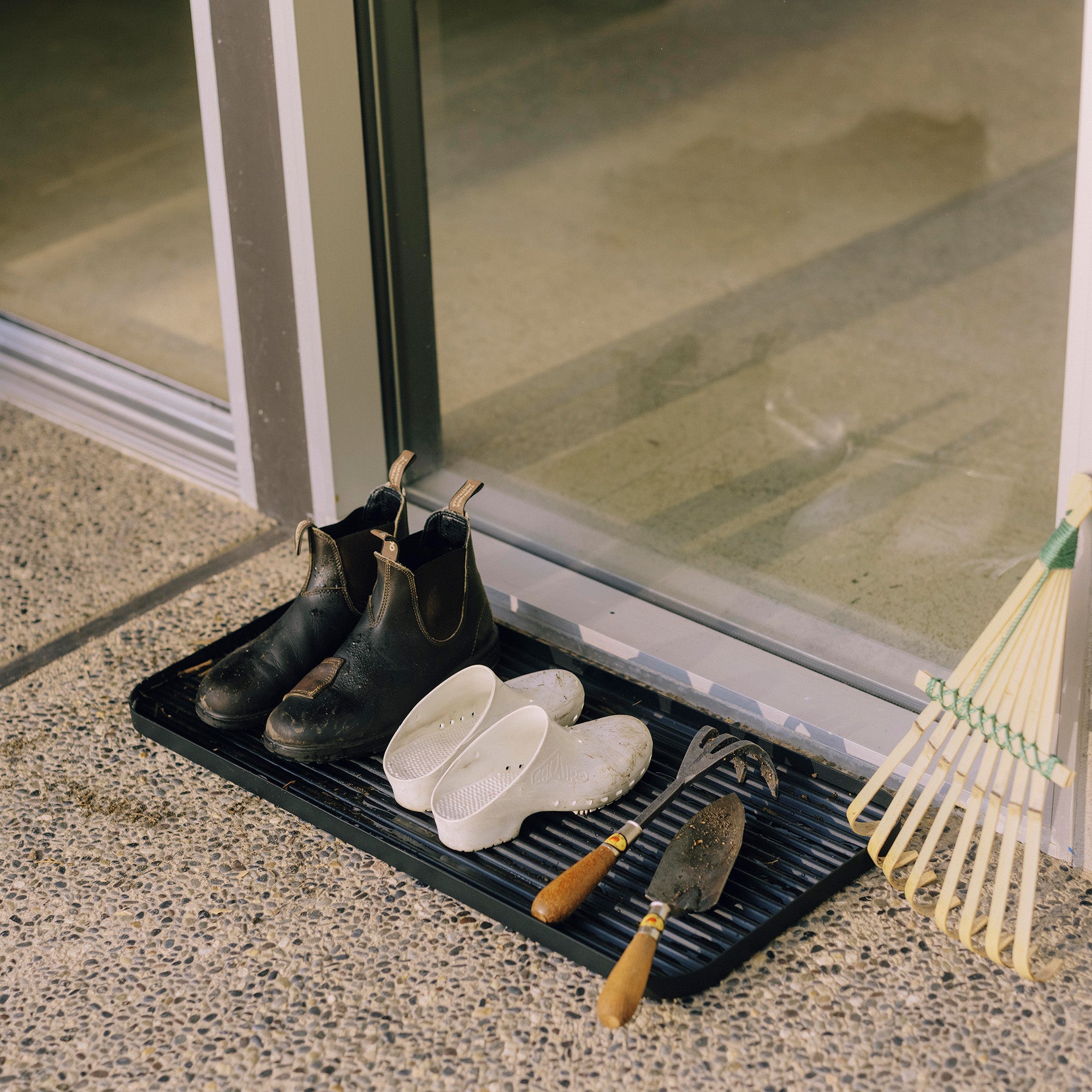Black steel boot tray from Apto outside a front door holding two pairs of shoes and gardening supplies.