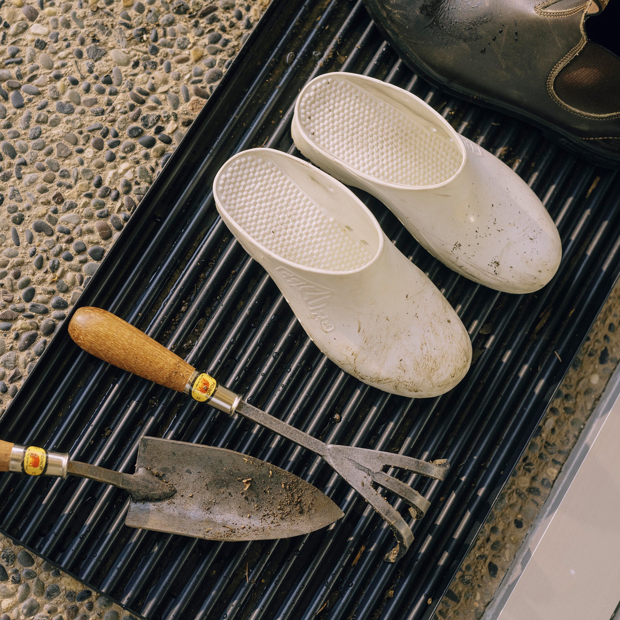 Black steel boot tray with ridged bottom holding two pairs of shoes and gardening supplies outside a modern home.