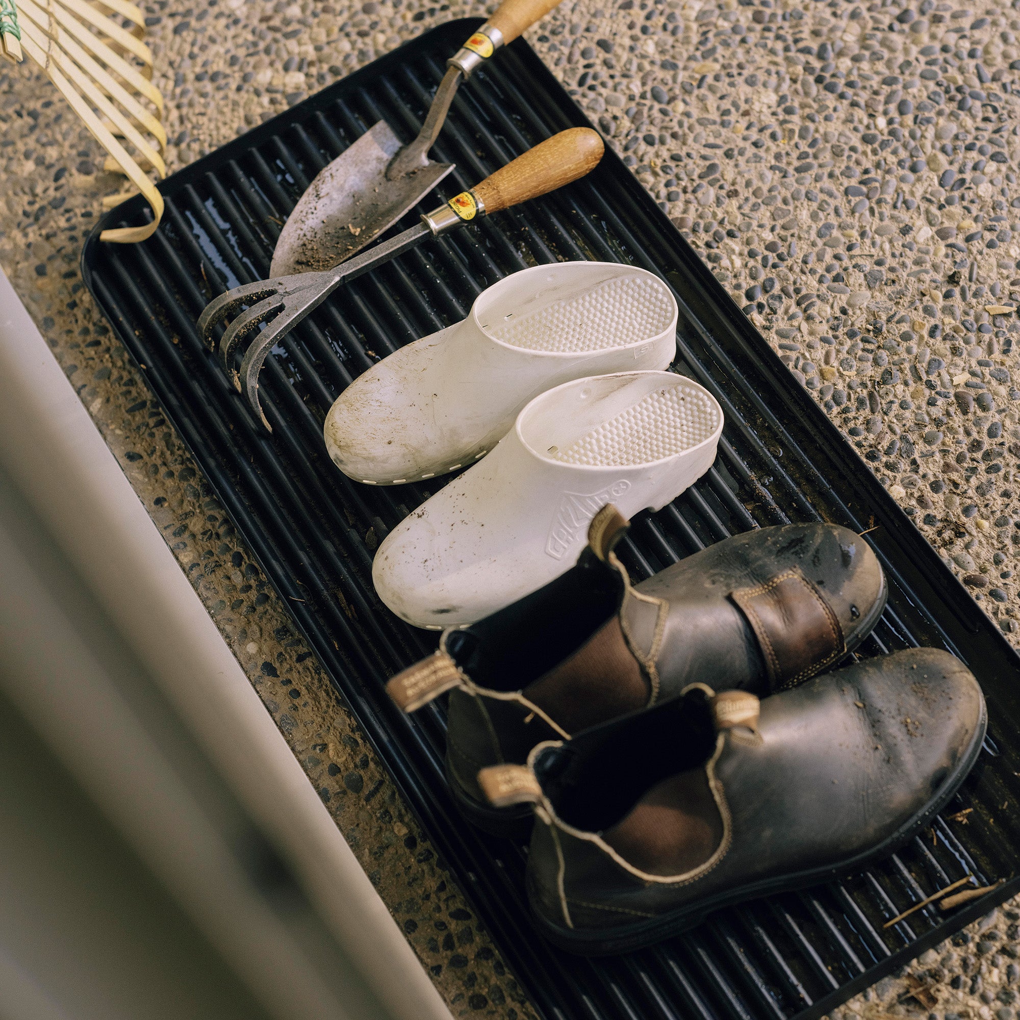 Black steel boot tray with ridged bottom holding a pair of gardening clogs, Blundstones and gardening supplies outside a modern home.