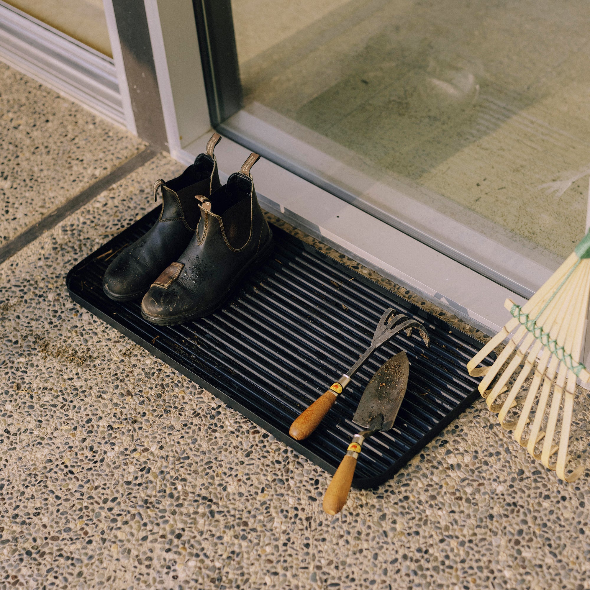 Black steel boot tray with ridged bottom holding a pair of Blundstones and gardening supplies outside a modern home.