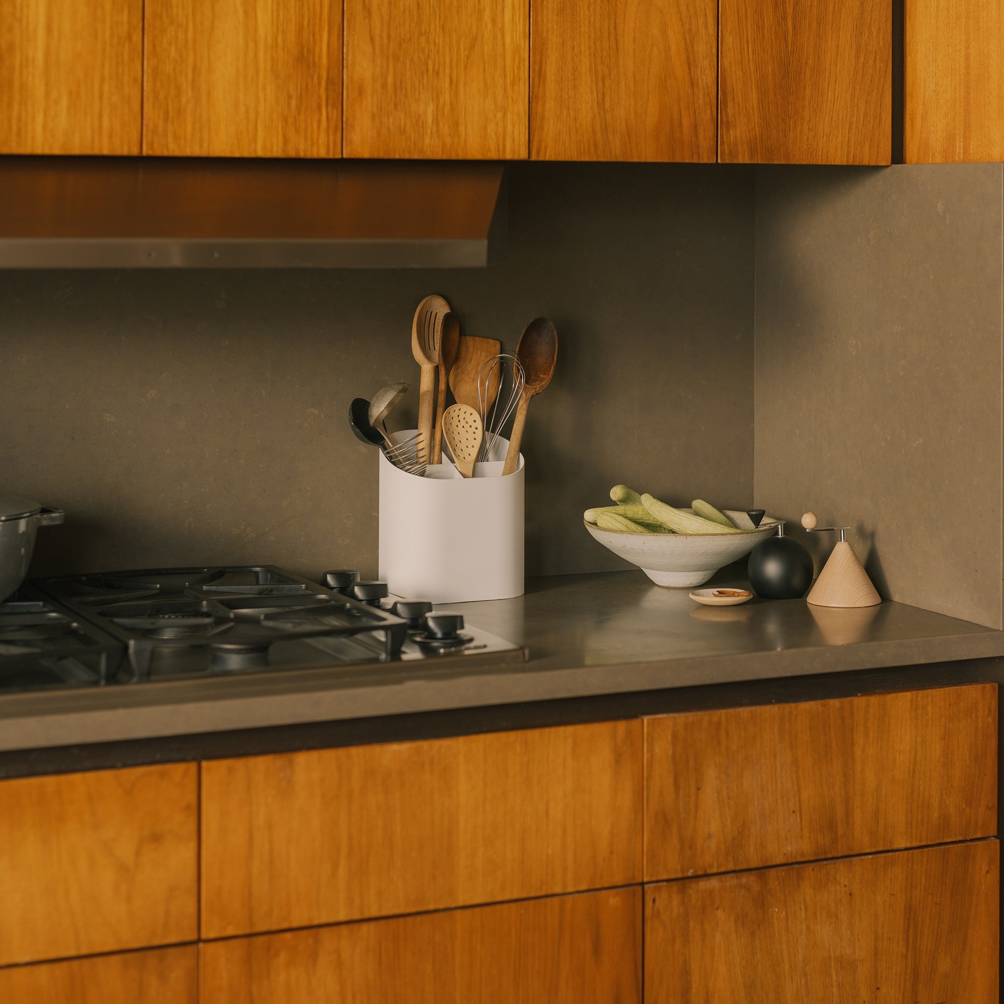 White kitchen utensil holder with wooden spoons on gray countertop beside gas stove and warm wood cabinets