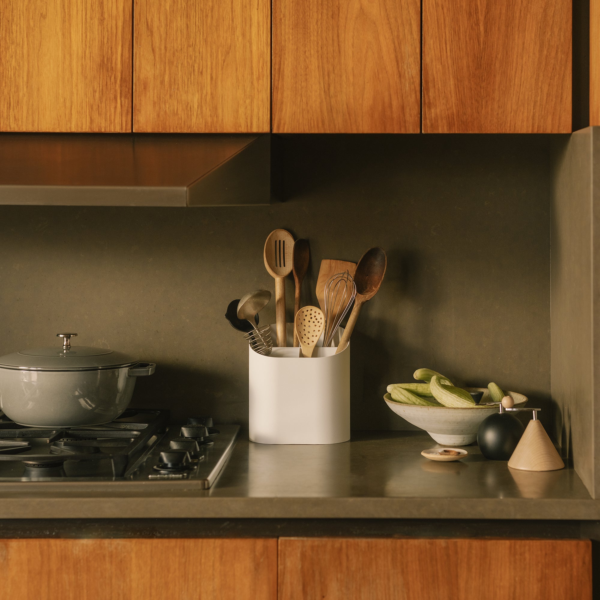 White kitchen utensil holder with sloped bottom holding wooden spoons on gray countertop beside gas stove and warm wood cabinets
