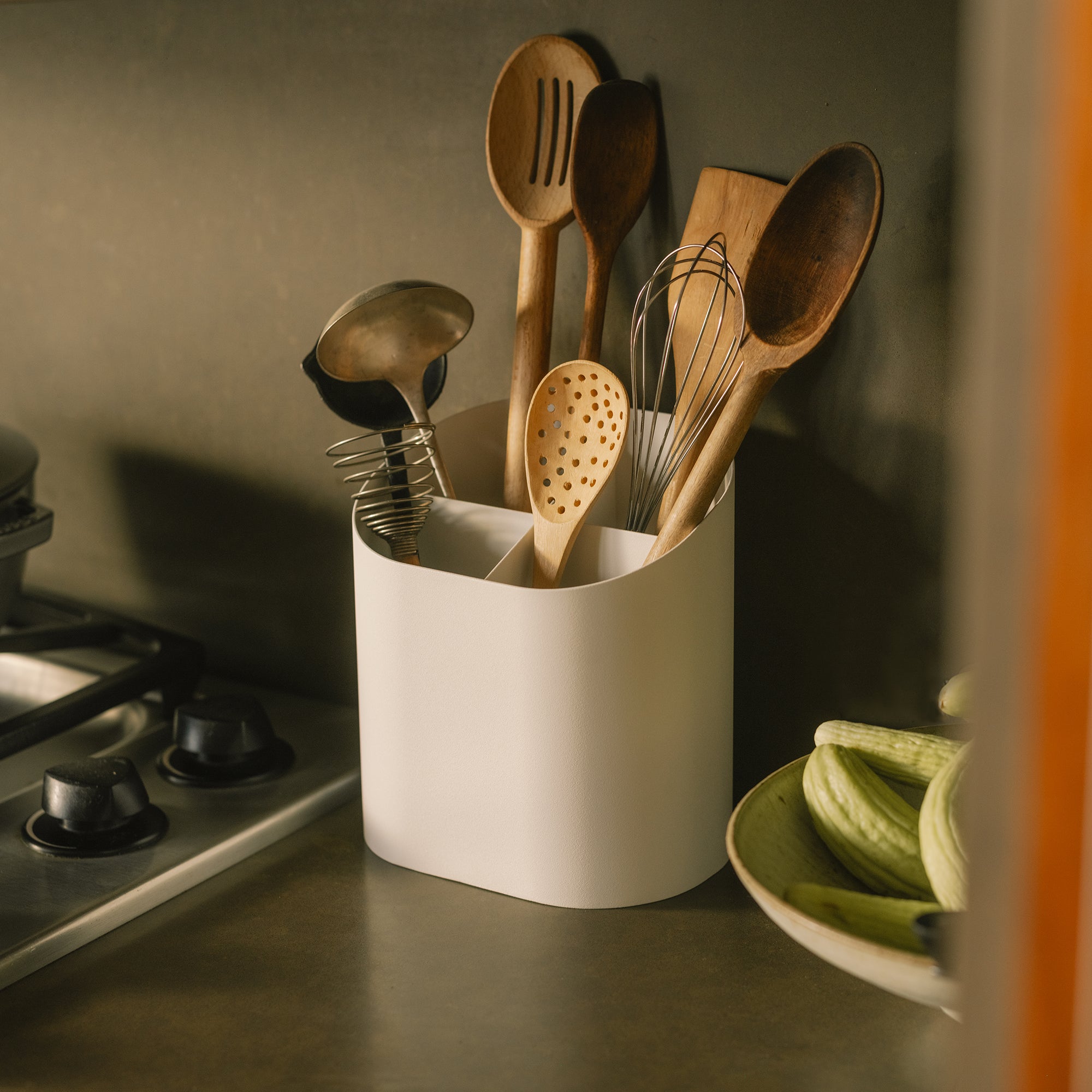 White utensil holder with divided compartments and sloped bottom storing wooden spoons and whisk next to gas stove