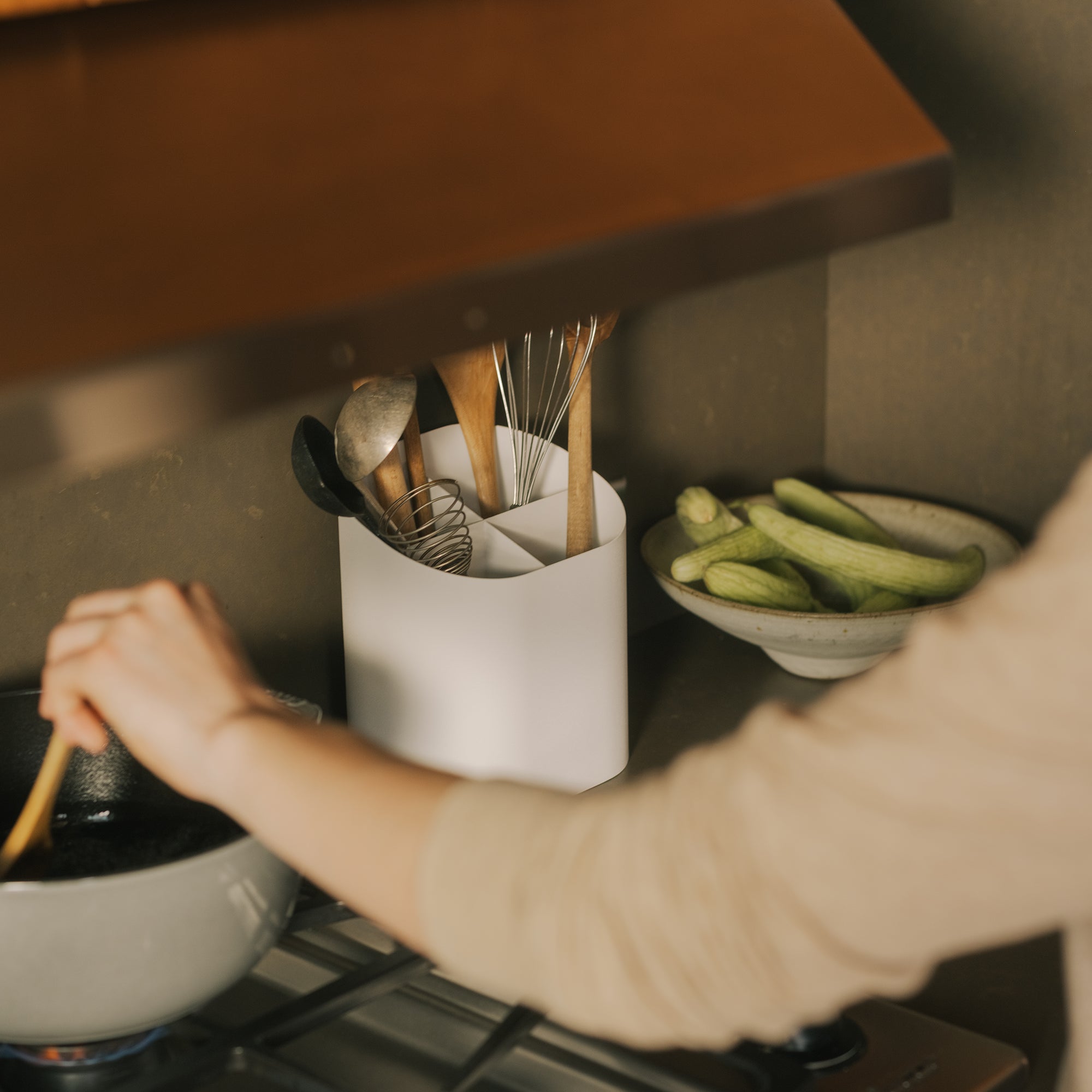 Person cooking on gas stove beside white utensil holder with wooden spoons and whisk on kitchen counter