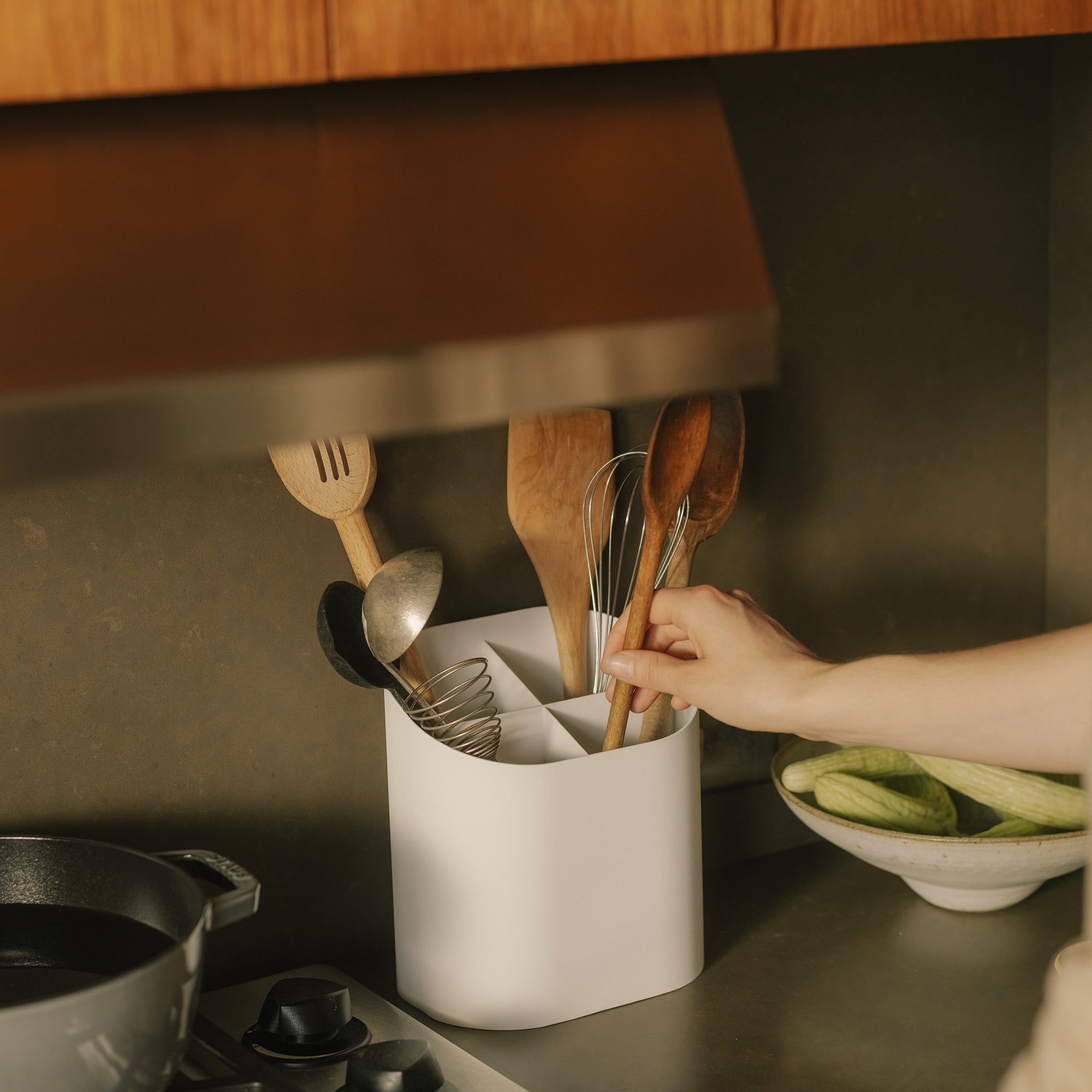 Hand reaching into white divided kitchen utensil holder with wooden spoons and whisk on countertop near stove