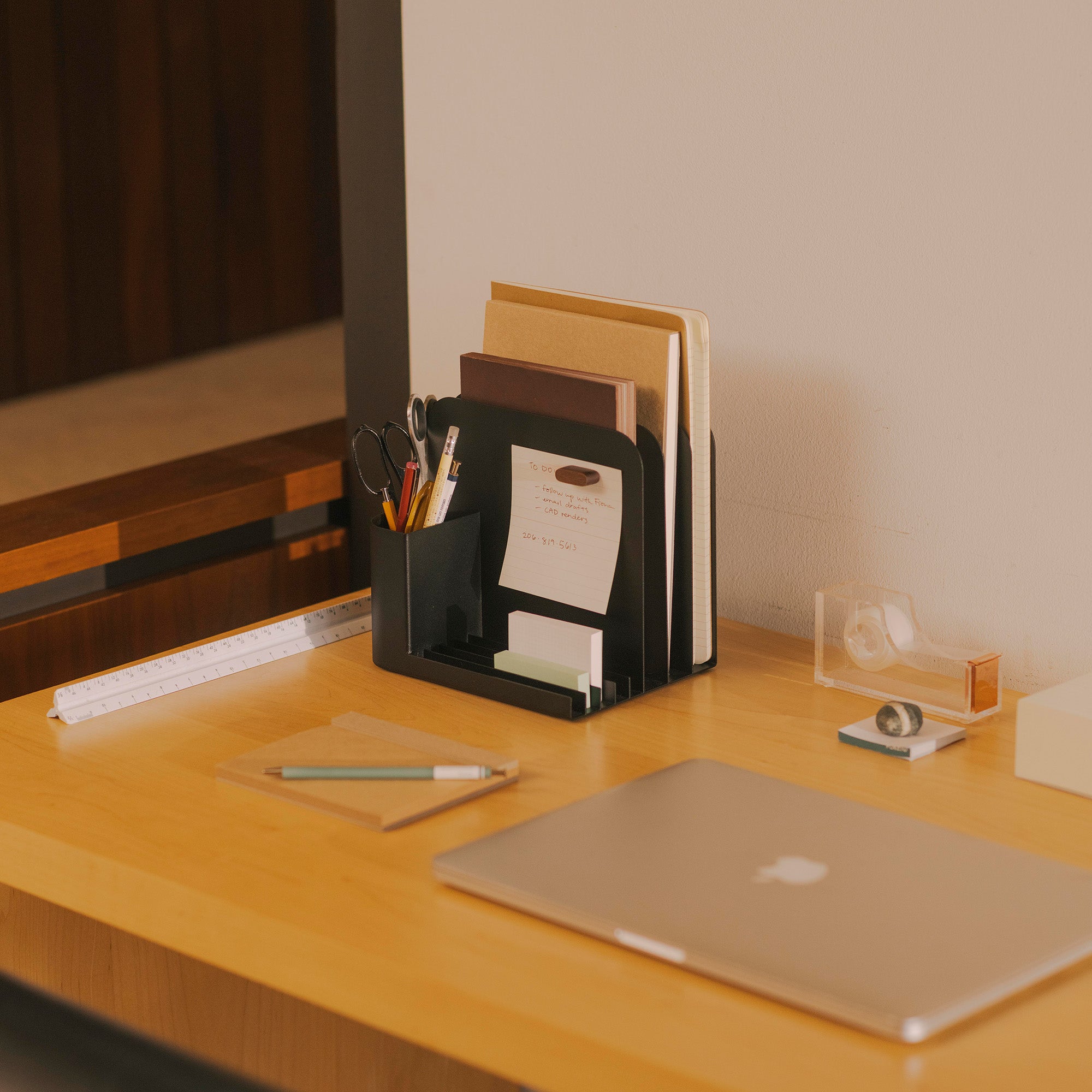 Black steel desk caddy with using slots for pens, files, and phones designed by Apto on a modern wood desk with a computer and other design tools. 