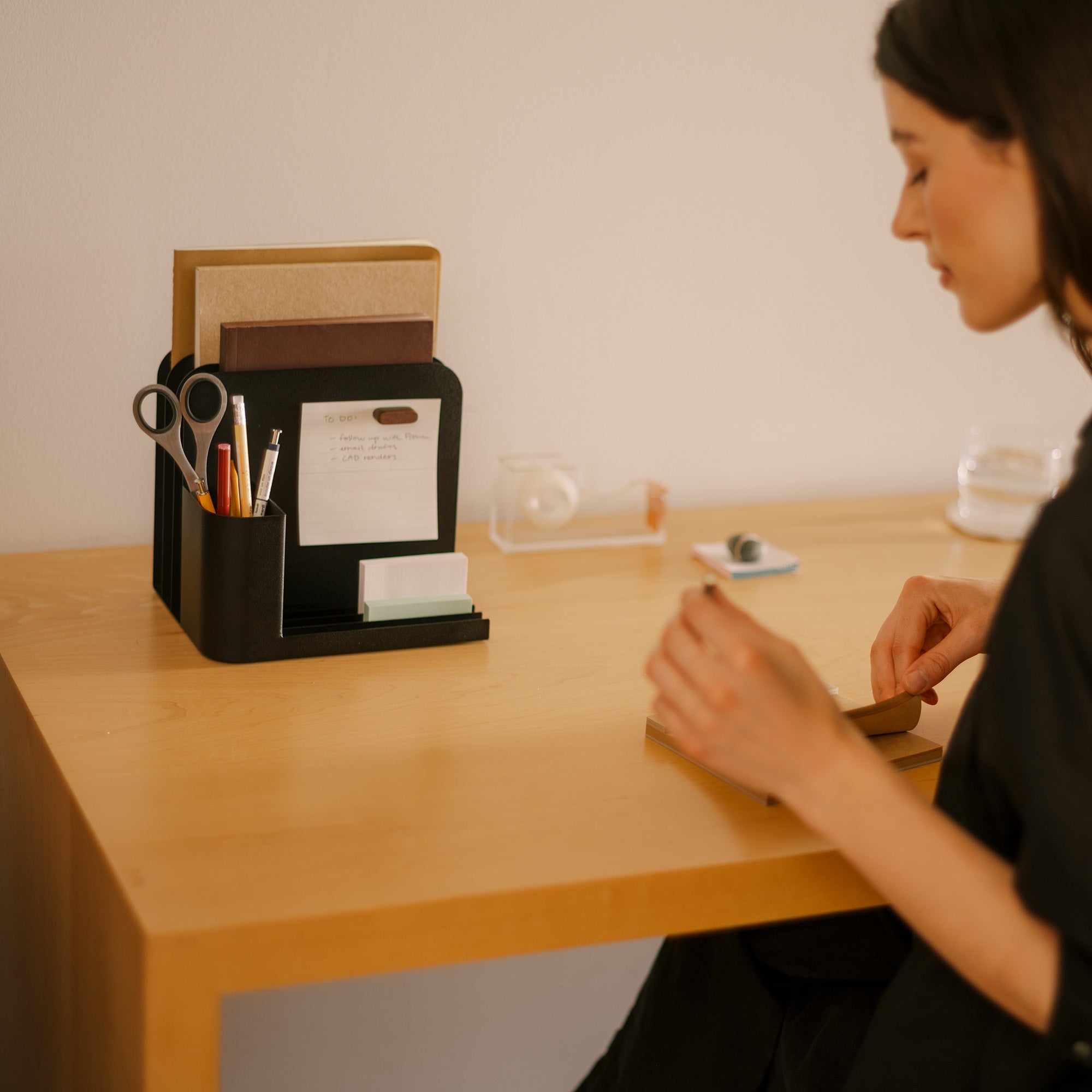 Woman works at a modern desk featuring a black steel desk caddy from Apto organizing files, pens, office supplies, and post-its.