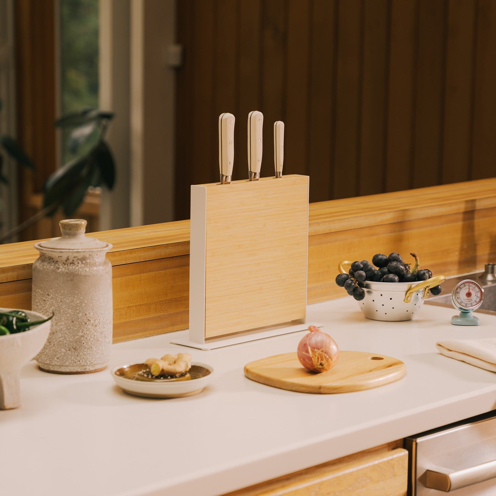 Apto Slot Knife Stand in bamboo and white steel holding three knives on a kitchen counter, next to a small round board with an apple