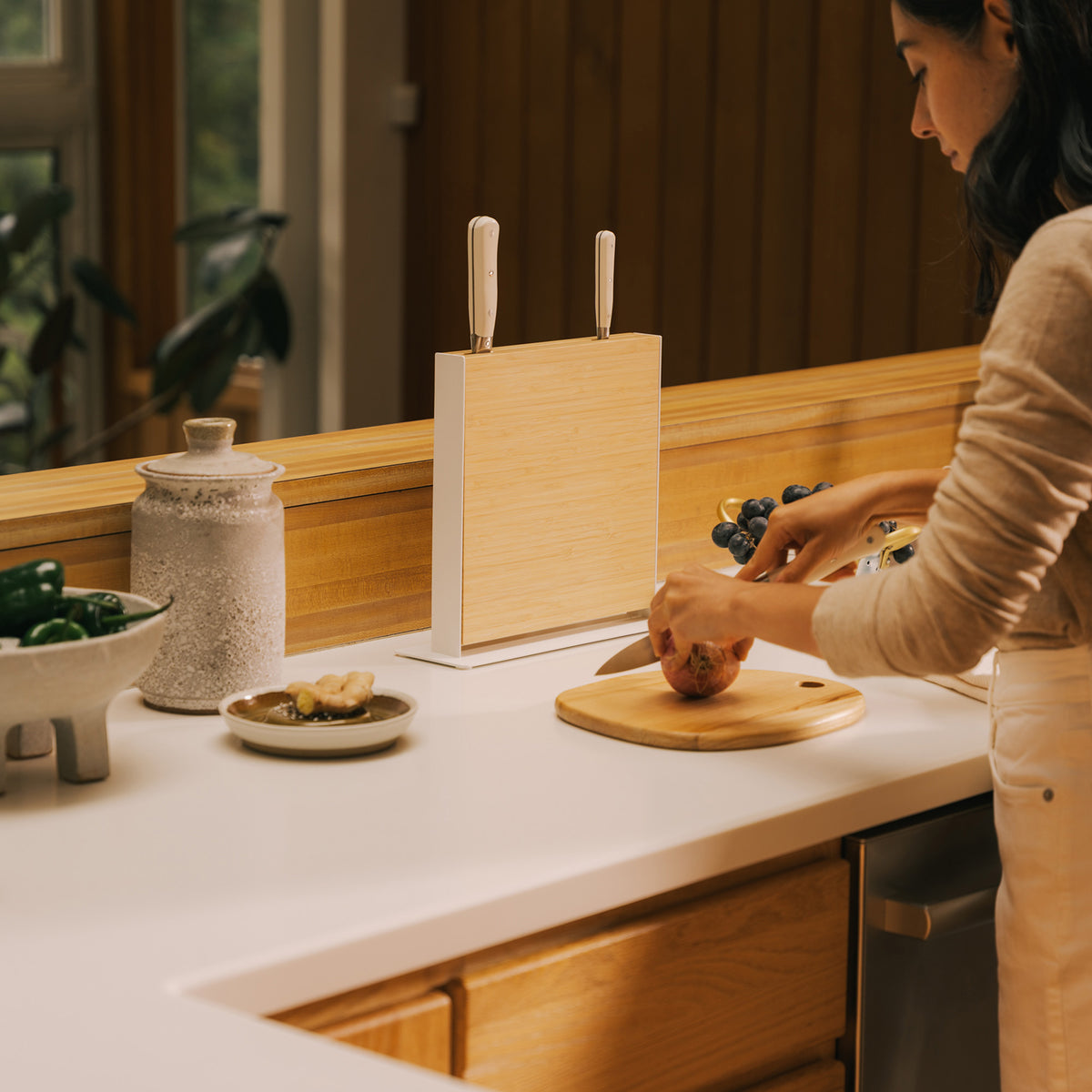 Woman chopping on a round board next to the Apto Slot Knife Stand in bamboo and white steel, which holds three knives on the counter