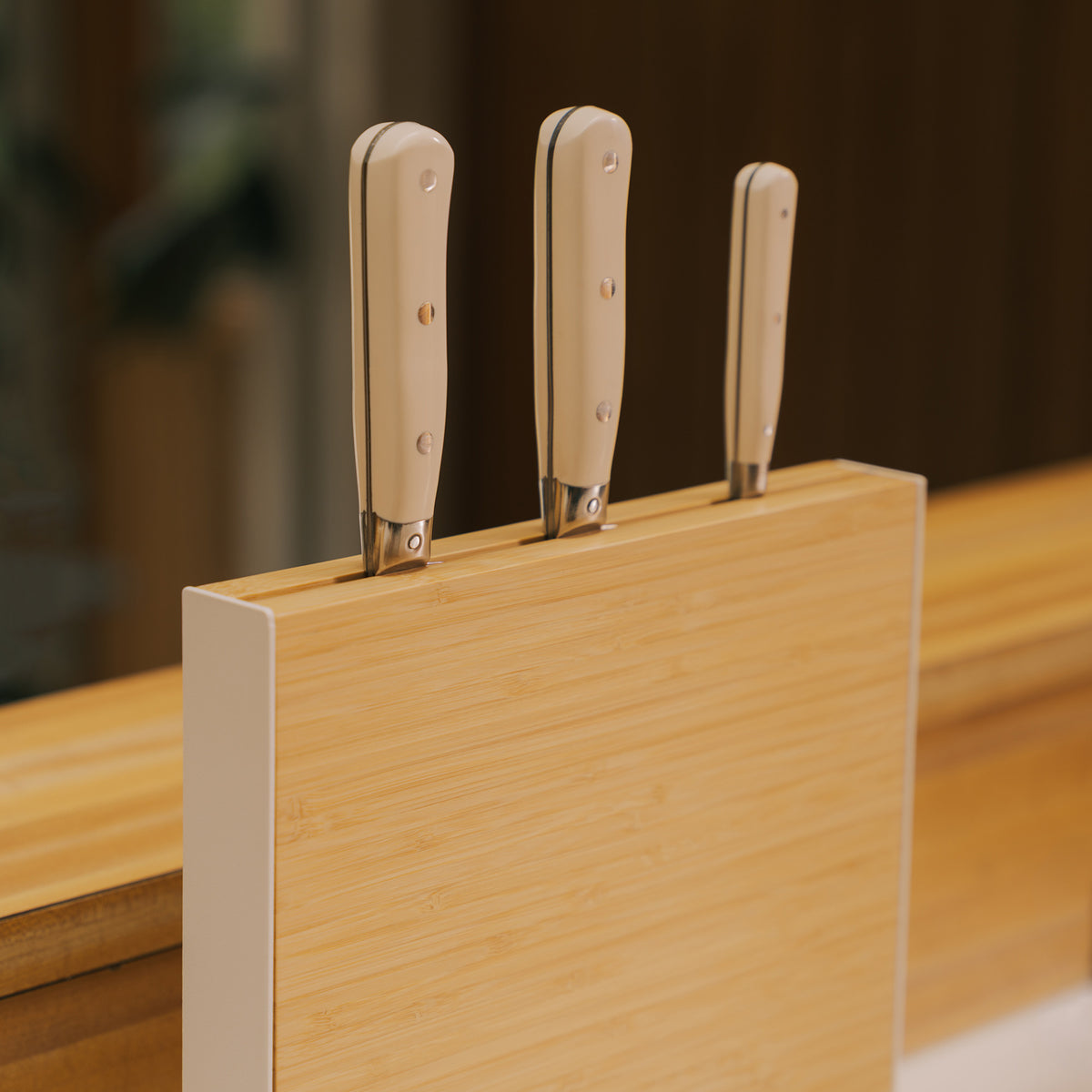 Close-up of three knives with light handles held upright in the Apto Slot Knife Stand, showing the bamboo panel and white steel frame