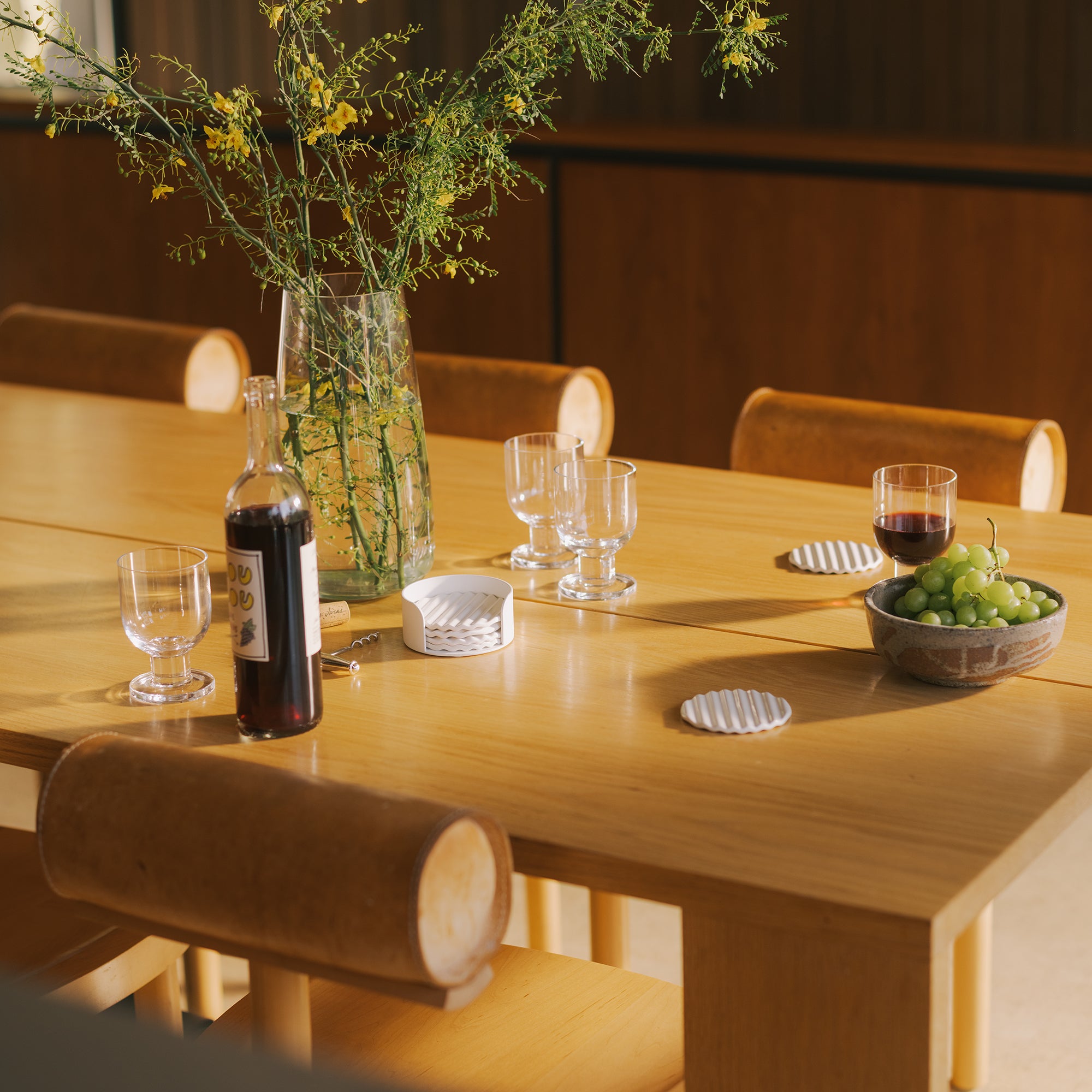 Oak dining table set with wine glasses, red wine bottle, white ceramic coasters in a set, grapes, and yellow wildflowers in warm natural light